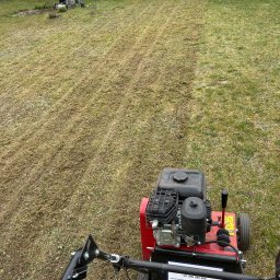 KO-OGRODY KAMIL POPIEL - Wertykulacja trawnika w ogrodzie. Widoczne ślady po wertykulatorze, osoba grabiąca trawę w tle, oraz sprzęt ogrodniczy i ławka. Ujęcie z perspektywy operatora wertykulatora.