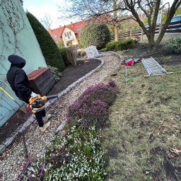 KO-OGRODY KAMIL POPIEL - Pracownik w czarnej bluzie używa dmuchawy do liści w ogrodzie z kwitnącymi wrzosami i kamienną ścieżką. Widoczne narzędzia ogrodnicze i drabina.