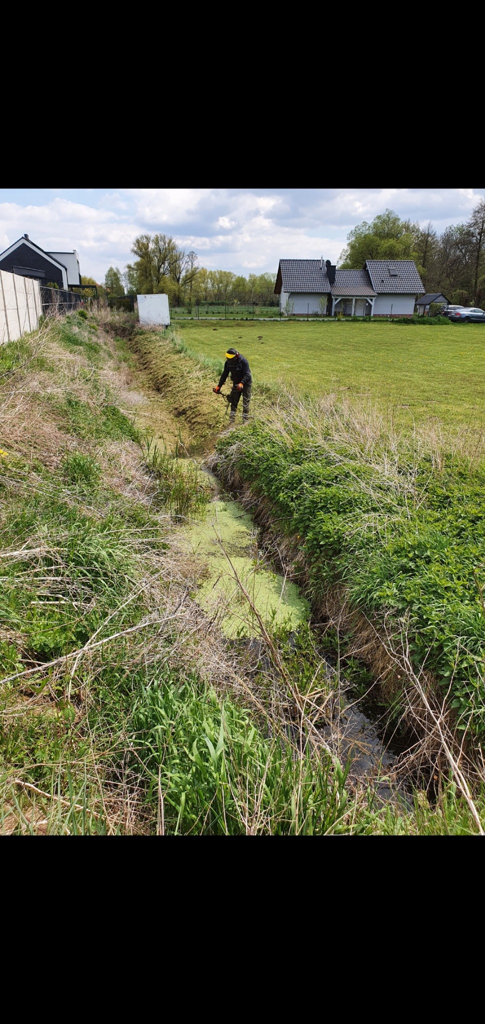 Mężczyzna w roboczym stroju i osłonie na twarzy przycina trawę w rowie melioracyjnym za pomocą podkaszarki spalinowej; widoczne domy w tle.