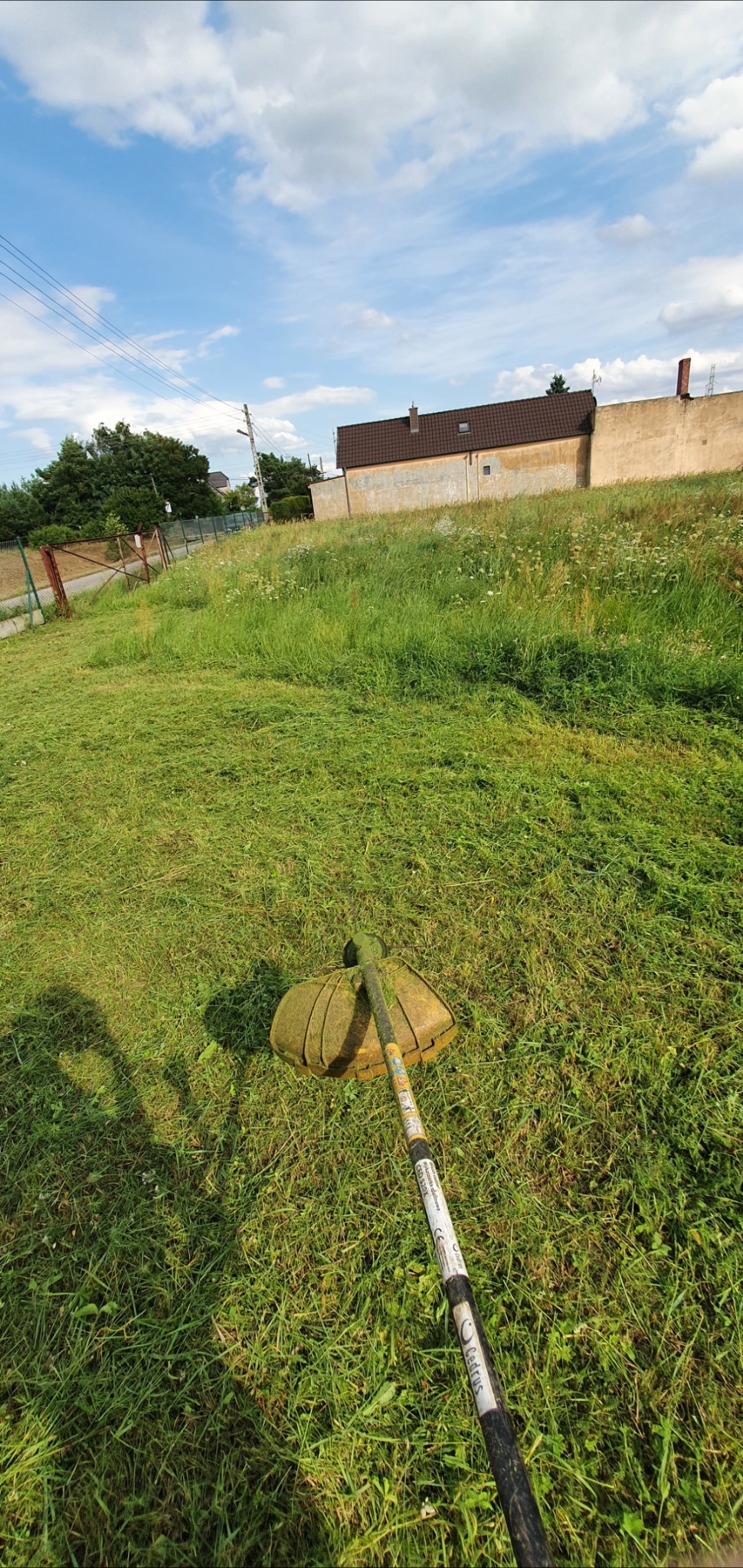 Widok z perspektywy pierwszej osoby na wykaszarkę spalinową podczas pracy na zarośniętym trawniku, z widocznym budynkiem w tle i błękitnym niebem z chmurami.