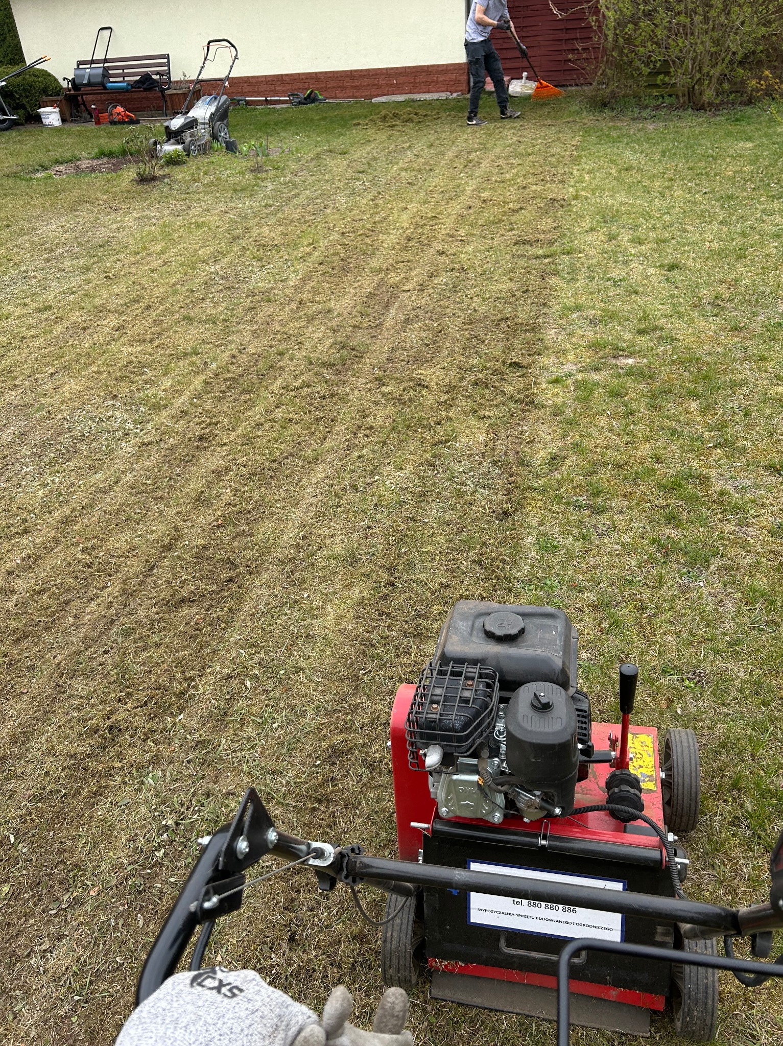 Wertykulacja trawnika w ogrodzie. Widoczne ślady po wertykulatorze, osoba grabiąca trawę w tle, oraz sprzęt ogrodniczy i ławka. Ujęcie z perspektywy operatora wertykulatora.