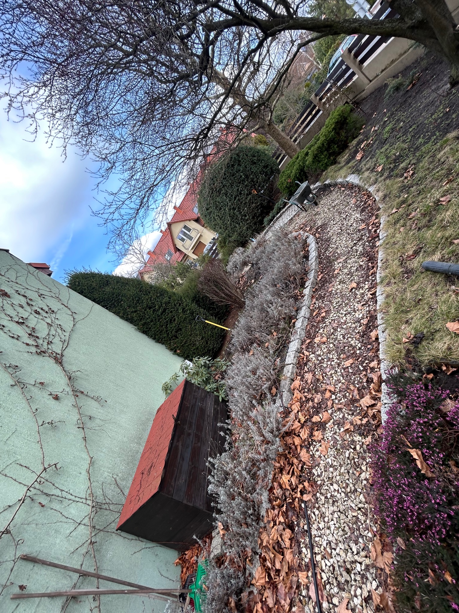 Kaskadowy ogród z kamienną ścieżką, obrzeżami i różnorodną roślinnością w jesiennej scenerii. Widoczny fragment budynku i niebo z chmurami.