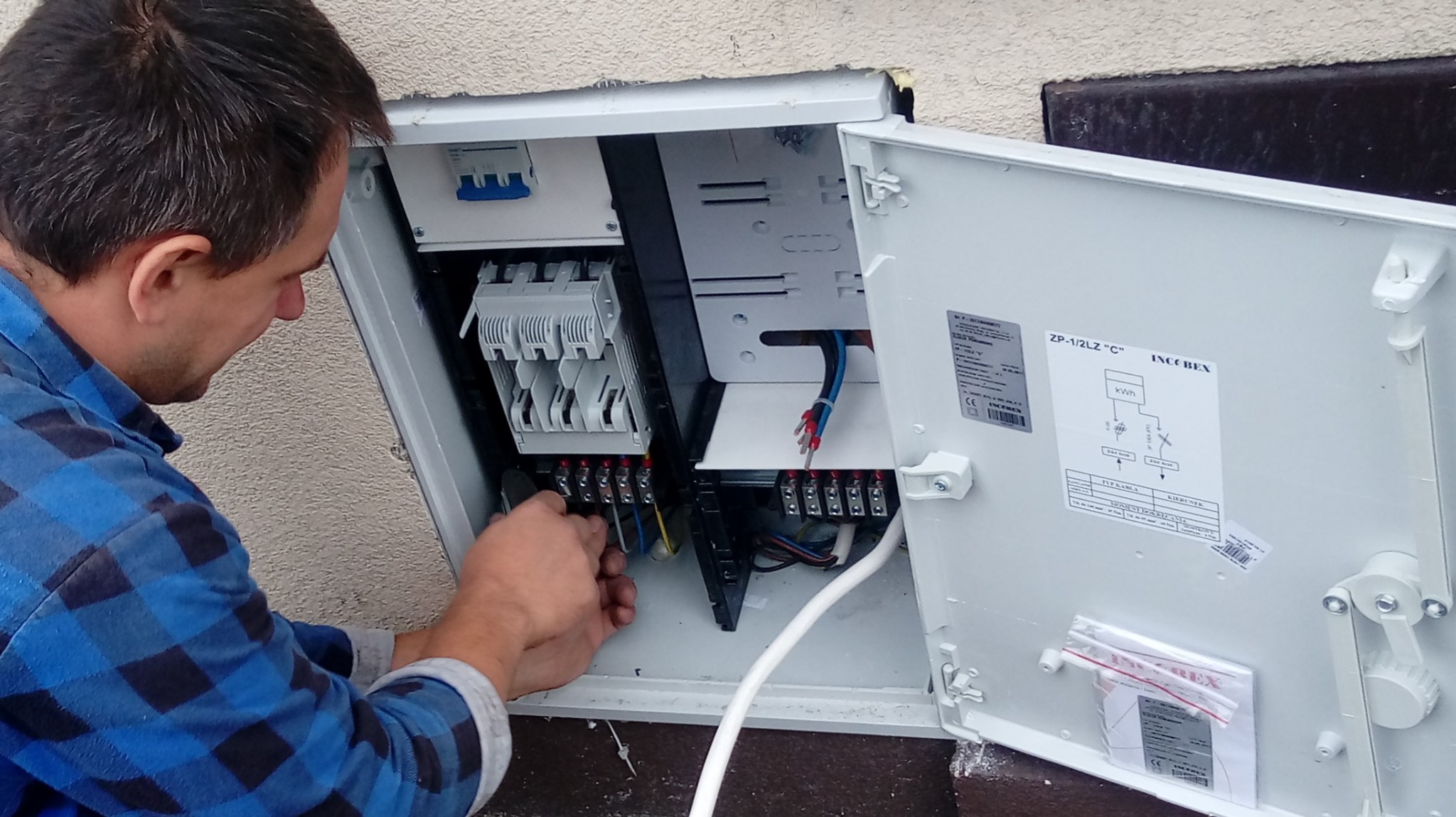 Elektryk w niebieskiej koszuli podłącza przewody w skrzynce elektrycznej zewnętrznej, widoczne bezpieczniki i schemat instalacji na drzwiczkach.