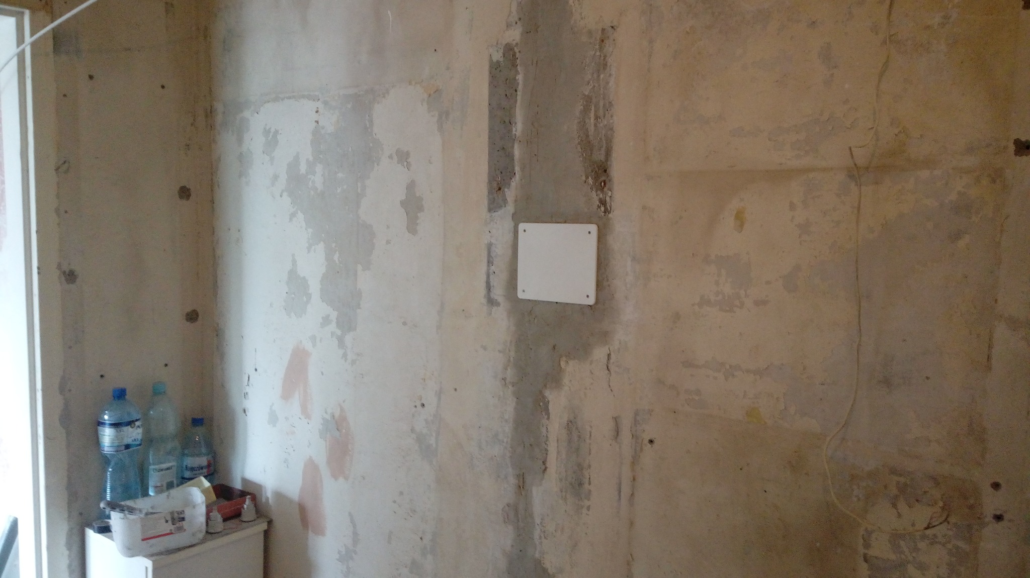 Nierówna ściana w trakcie remontu z widoczną puszką instalacyjną, pozostałościami farby i butelkami wody na podłodze.