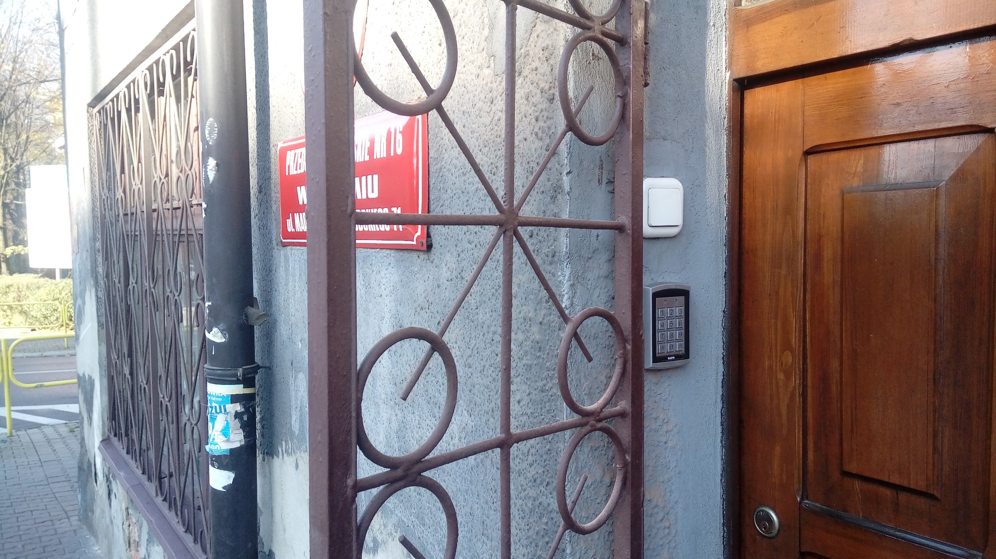Zewnętrzny panel domofonowy z klawiaturą numeryczną, zamontowany obok brązowych drzwi wejściowych i ozdobnej metalowej kraty