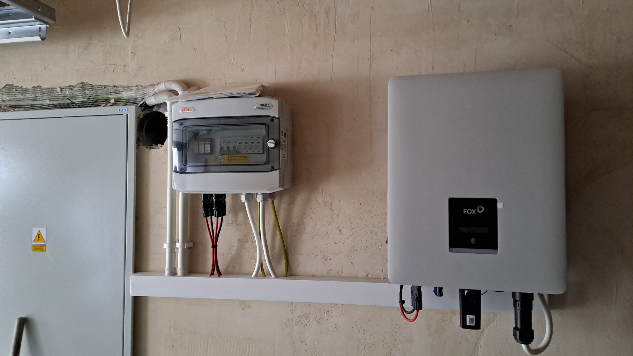 Szara skrzynka elektryczna z bezpiecznikami i przewodami oraz biały inwerter firmy FOX zamontowane na ścianie o nierównej fakturze, widoczny otwór w ścianie z kablami.