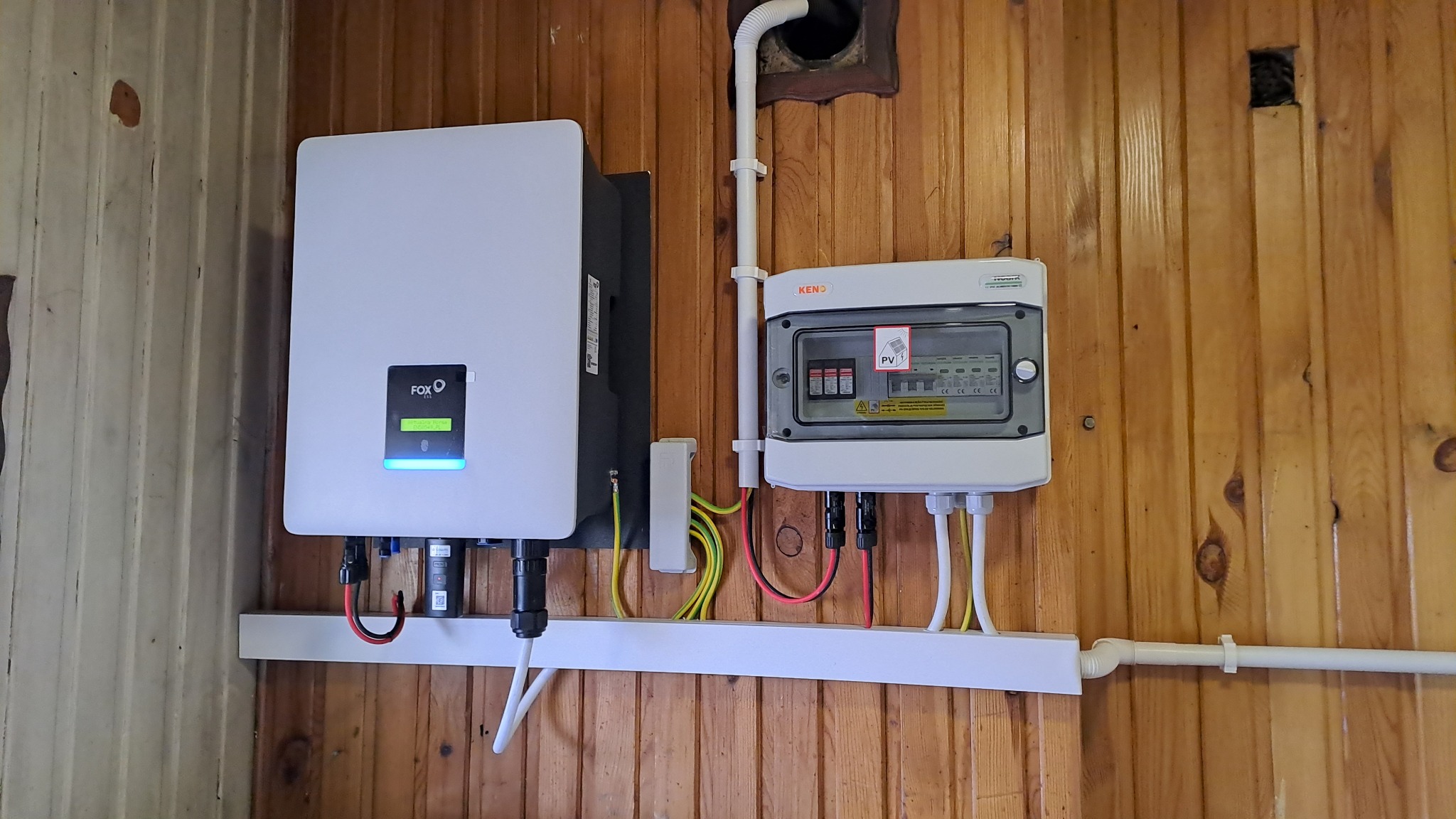 Biały falownik fotowoltaiczny FoxESS i skrzynka zabezpieczeń KENO zamontowane na drewnianej ścianie z widocznymi przewodami i rurami instalacyjnymi.