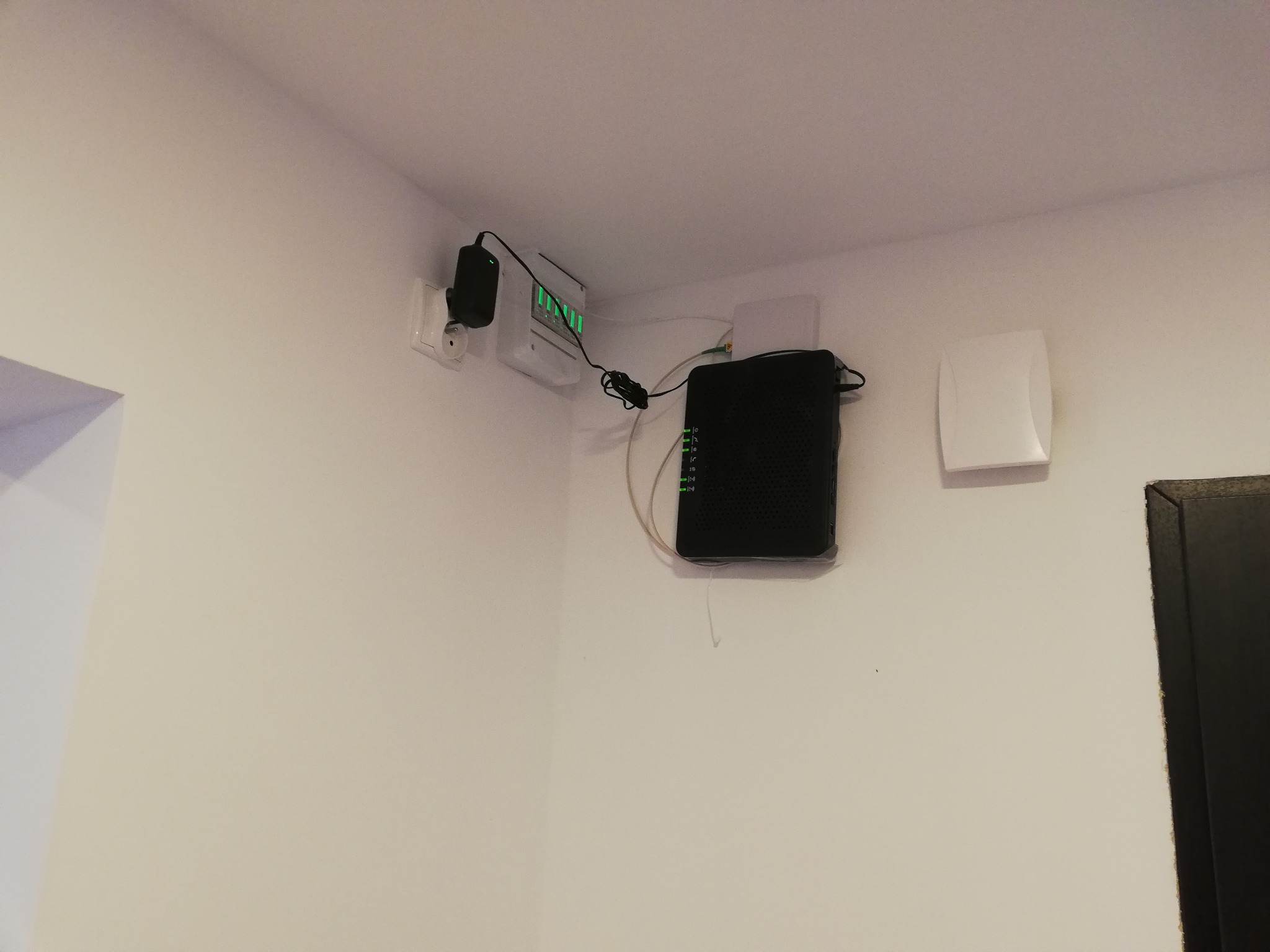 Rozdzielnia elektryczna z zielonymi diodami, router z podłączonymi kablami oraz wentylator zamontowane na białej ścianie w rogu pomieszczenia.