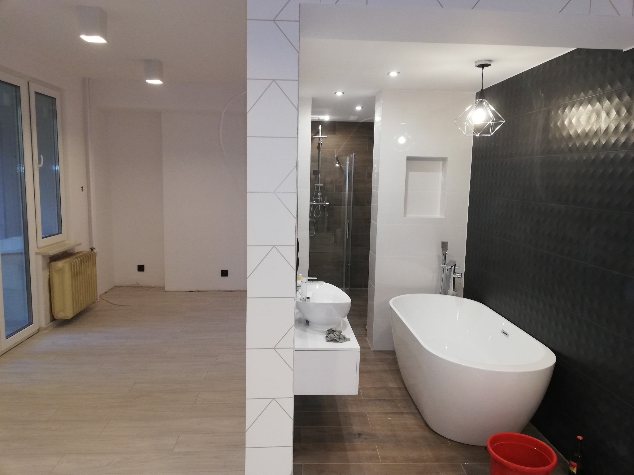 Wnętrze łazienki z wolnostojącą białą wanną, umywalką nablatową i kabiną prysznicową, widoczne przez otwór w ścianie z dekoracyjnymi płytkami, w tle pokój z grzejnikiem i oknem balkonowym.