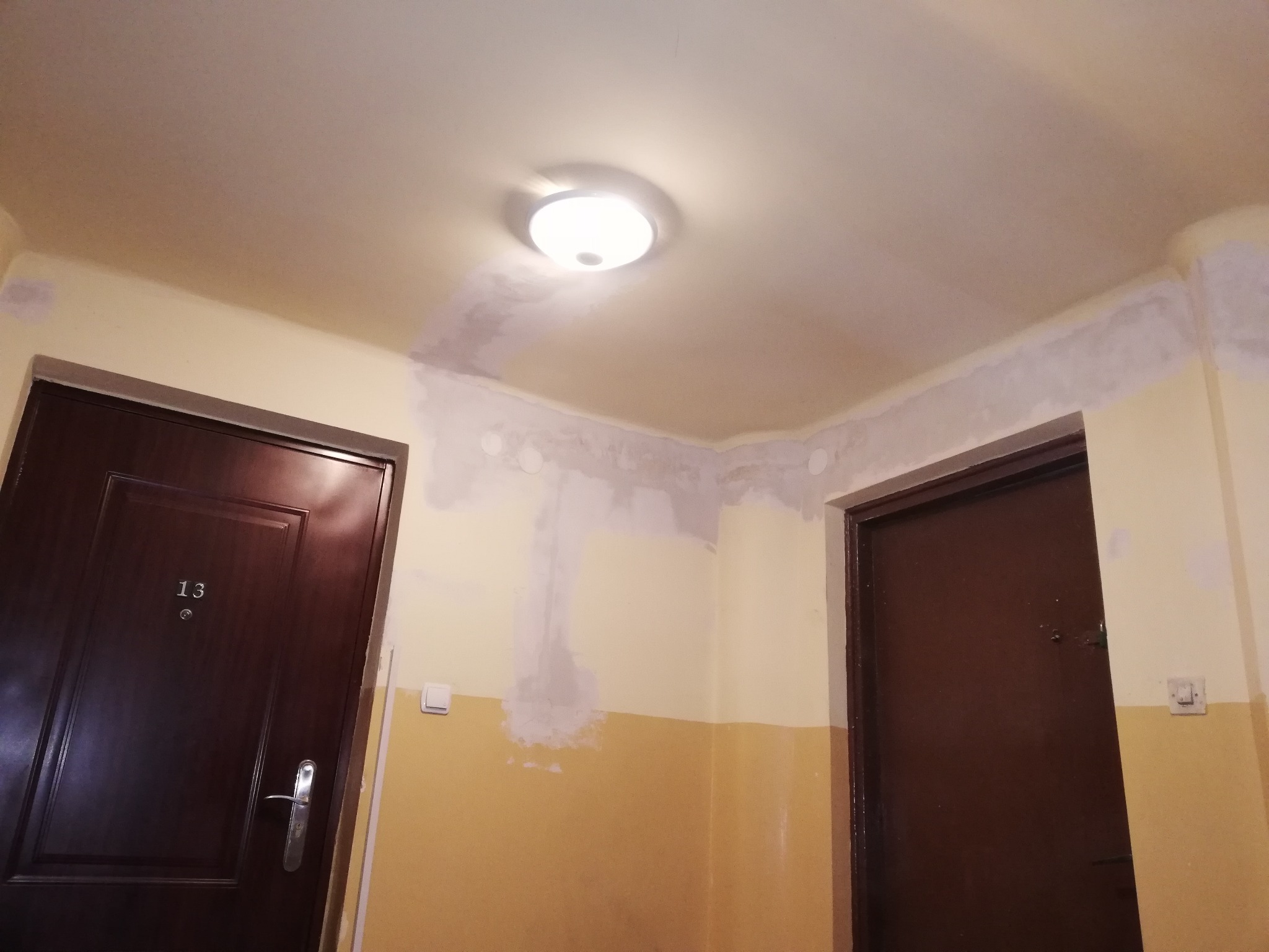 Korytarz w bloku po częściowym malowaniu ścian i sufitu, widoczne ślady po instalacjach elektrycznych i gniazdach, dwie brązowe drzwi wejściowe do mieszkań, numer 13 na jednych z nich.