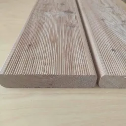 Dwie deski tarasowe z ryflowaną powierzchnią, widoczne słoje drewna i zaokrąglone krawędzie, ułożone na drewnianym blacie.