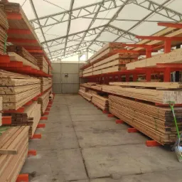 Magazyn z rzędami tarcicy ułożonej na pomarańczowych wspornikach, widoczna szara podłoga i metalowe drzwi w tle, oświetlony przez białe zadaszenie z metalową konstrukcją.