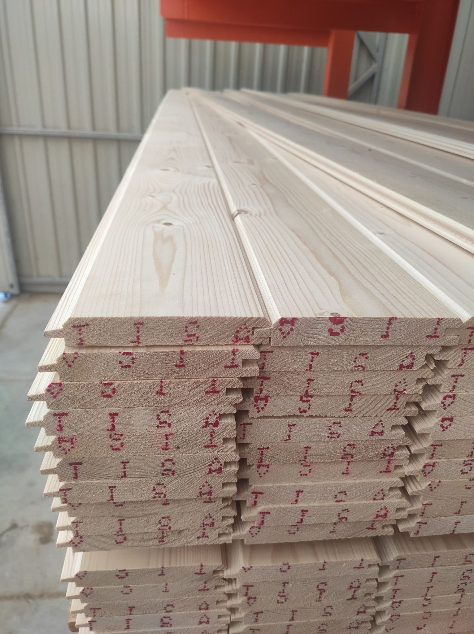 Stos jasnej, drewnianej boazerii z widocznymi czerwonymi oznaczeniami na krawędziach, ułożonej w magazynie z metalową konstrukcją w tle.