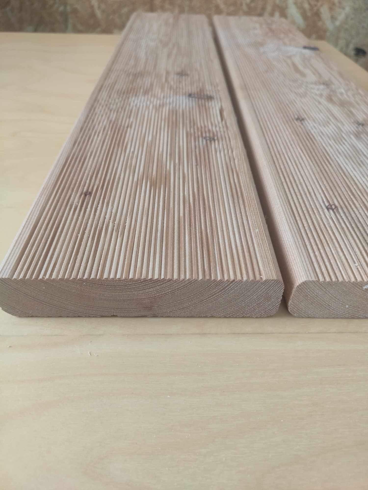 Dwie deski tarasowe z ryflowaną powierzchnią, widoczne słoje drewna i zaokrąglone krawędzie, ułożone na drewnianym blacie.