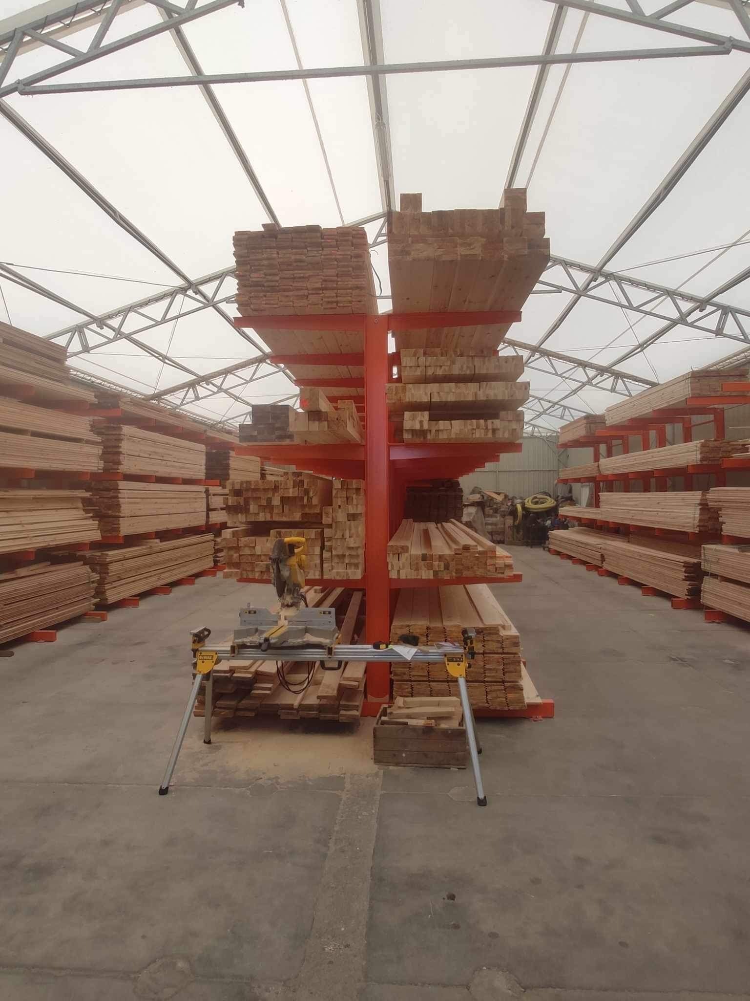 Magazyn tarcicy w hali z metalową konstrukcją i białym dachem; na pierwszym planie piła tarczowa na stojaku, w tle rzędy ułożonej tarcicy na pomarańczowych regałach.