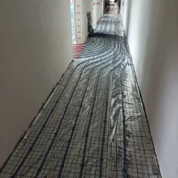 Instalacja ogrzewania podłogowego w korytarzu, rurki w kolorze niebieskim ułożone na siatce z folią odblaskową, widoczne kable pod sufitem.