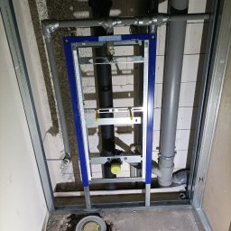 K.G Instal - Stelaż podtynkowy WC zamontowany w łazience w trakcie remontu, widoczne rury kanalizacyjne i odpływ podłogowy, ściany w surowym stanie.