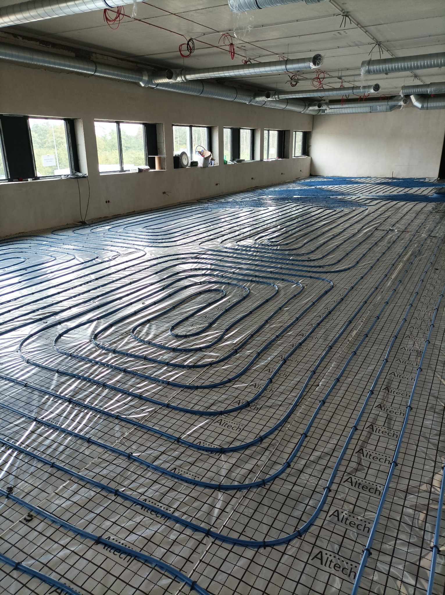 Niebieskie rury ogrzewania podłogowego ułożone na izolacji z nadrukowaną siatką w surowym pomieszczeniu z oknami i instalacją wentylacyjną pod sufitem.