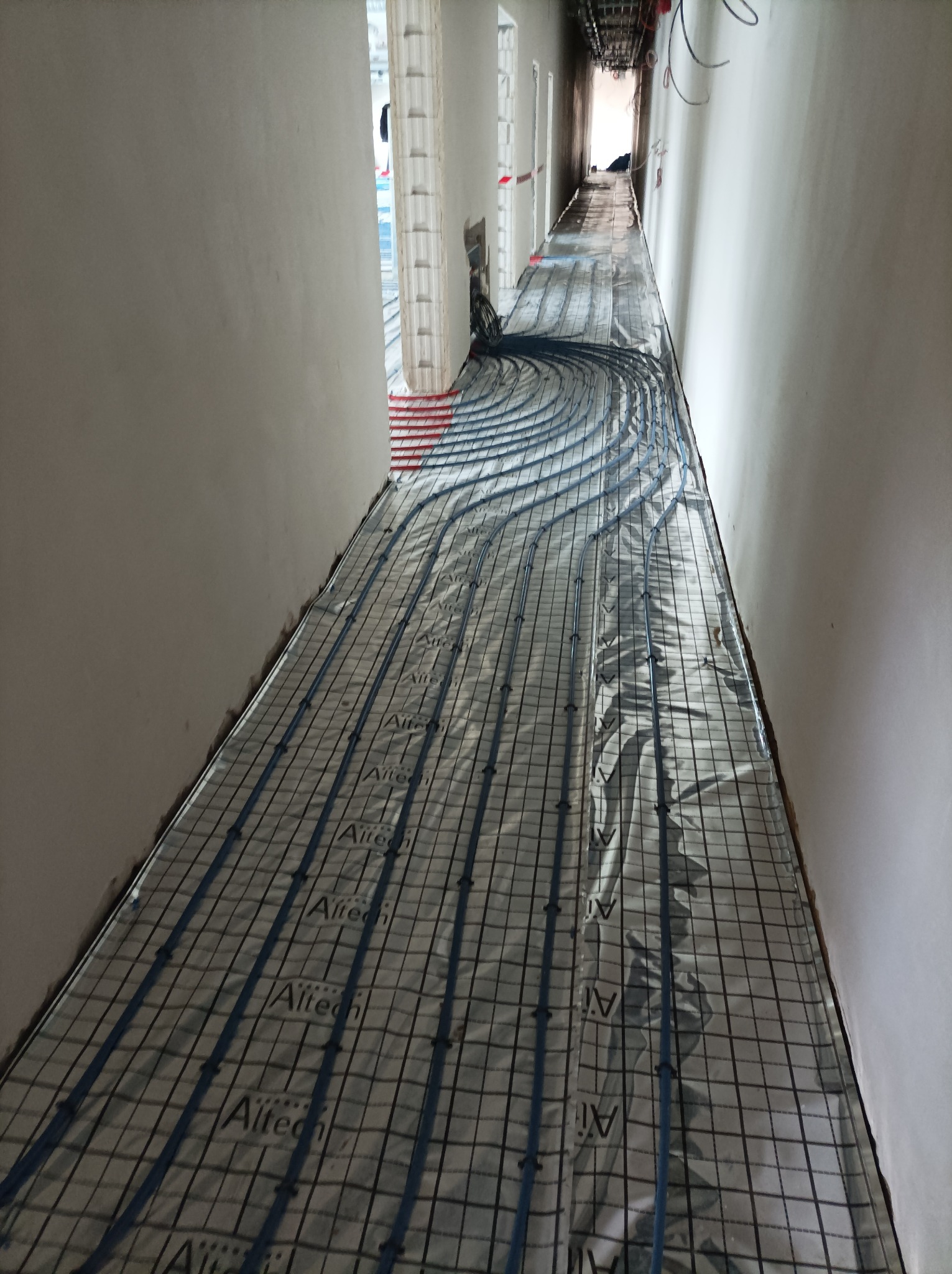 Instalacja ogrzewania podłogowego w korytarzu, rurki w kolorze niebieskim ułożone na siatce z folią odblaskową, widoczne kable pod sufitem.