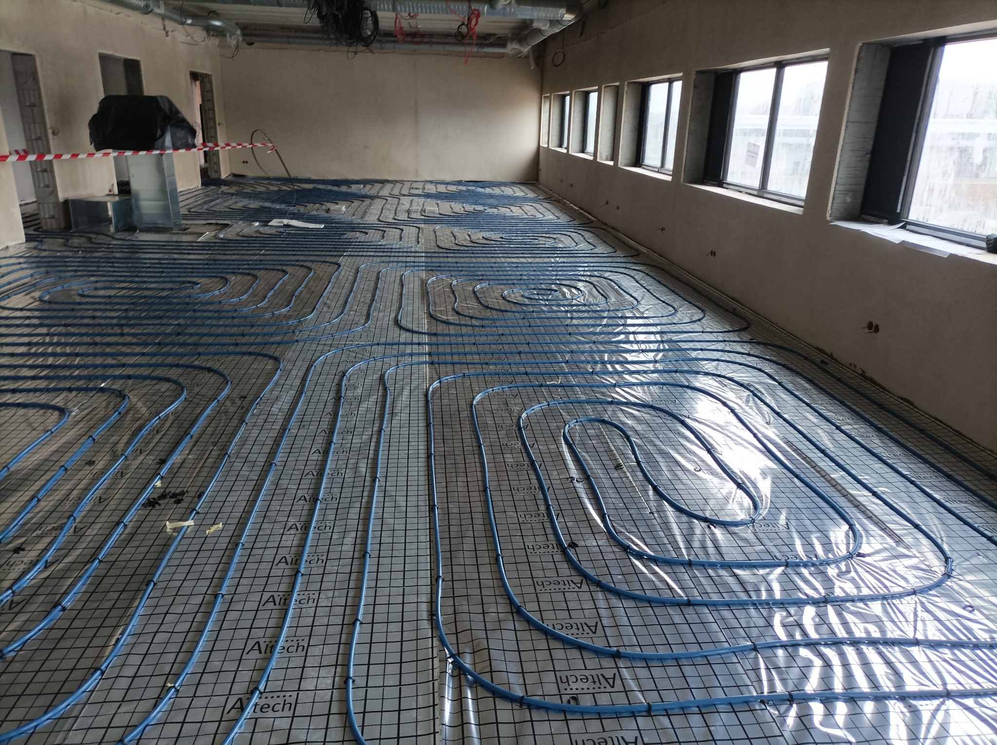 Niebieskie rury ogrzewania podłogowego ułożone na siatce z folią aluminiową w surowym wnętrzu budynku w trakcie instalacji. Widoczne okna i elementy instalacji pod sufitem.