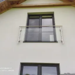 Szklana balustrada balkonowa z nierdzewnymi elementami mocującymi, zamontowana na elewacji domu z widocznym drewnianym okapem dachu.