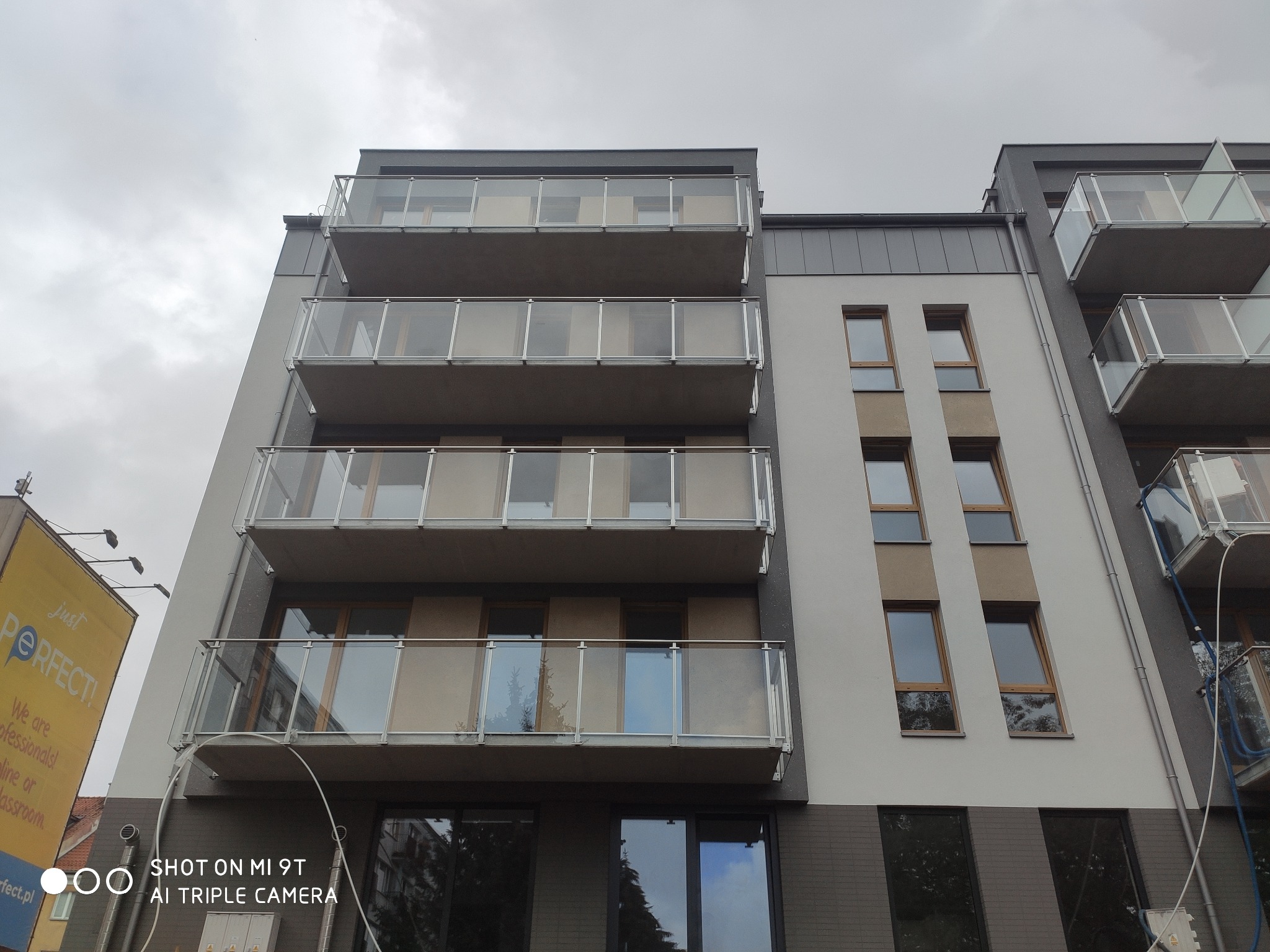 Szklane balustrady balkonowe w nowoczesnym budynku mieszkalnym, widok z dołu na kilka pięter, pochmurne niebo w tle.