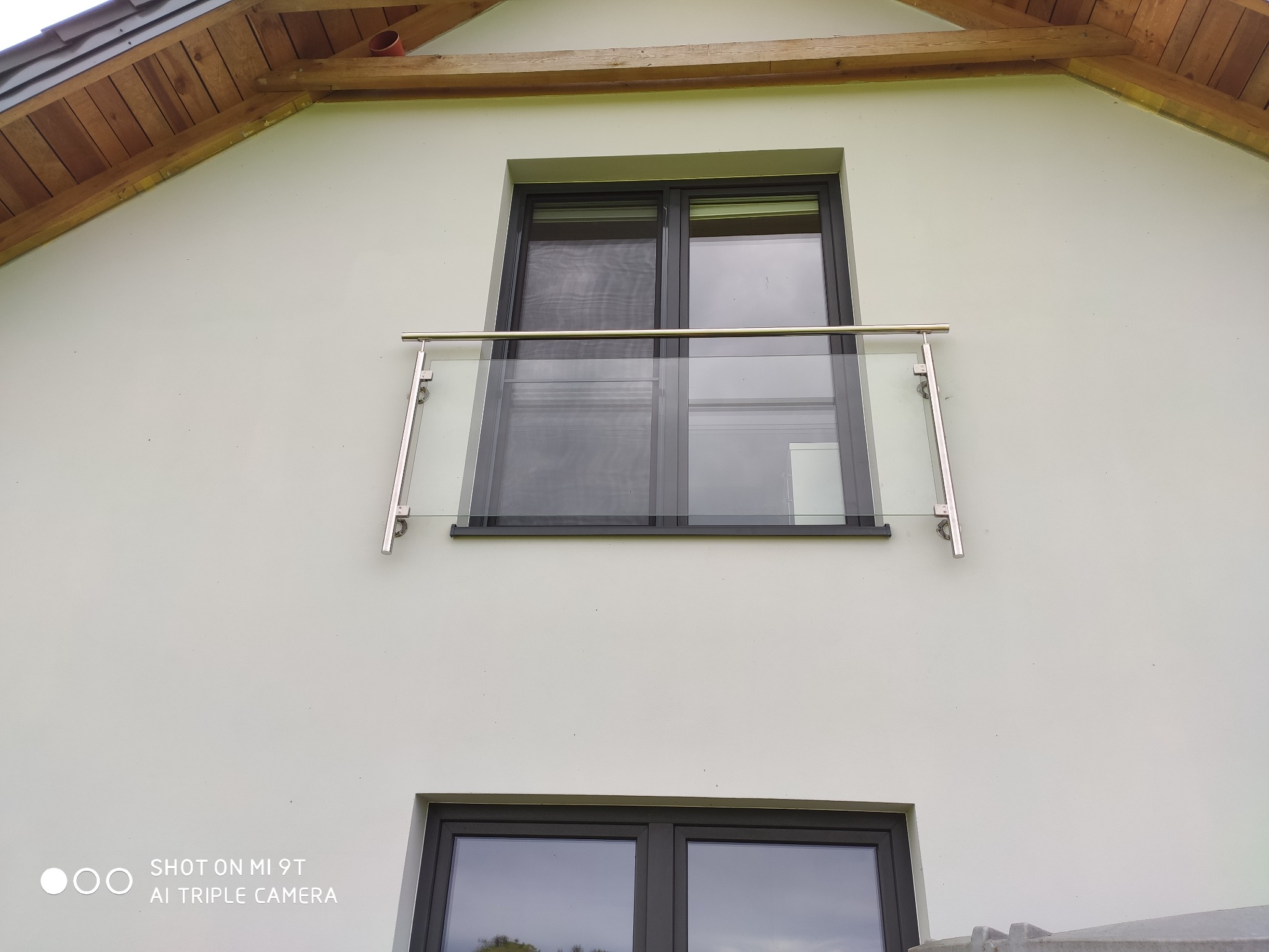 Szklana balustrada balkonowa z nierdzewnymi elementami mocującymi, zamontowana na elewacji domu z widocznym drewnianym okapem dachu.