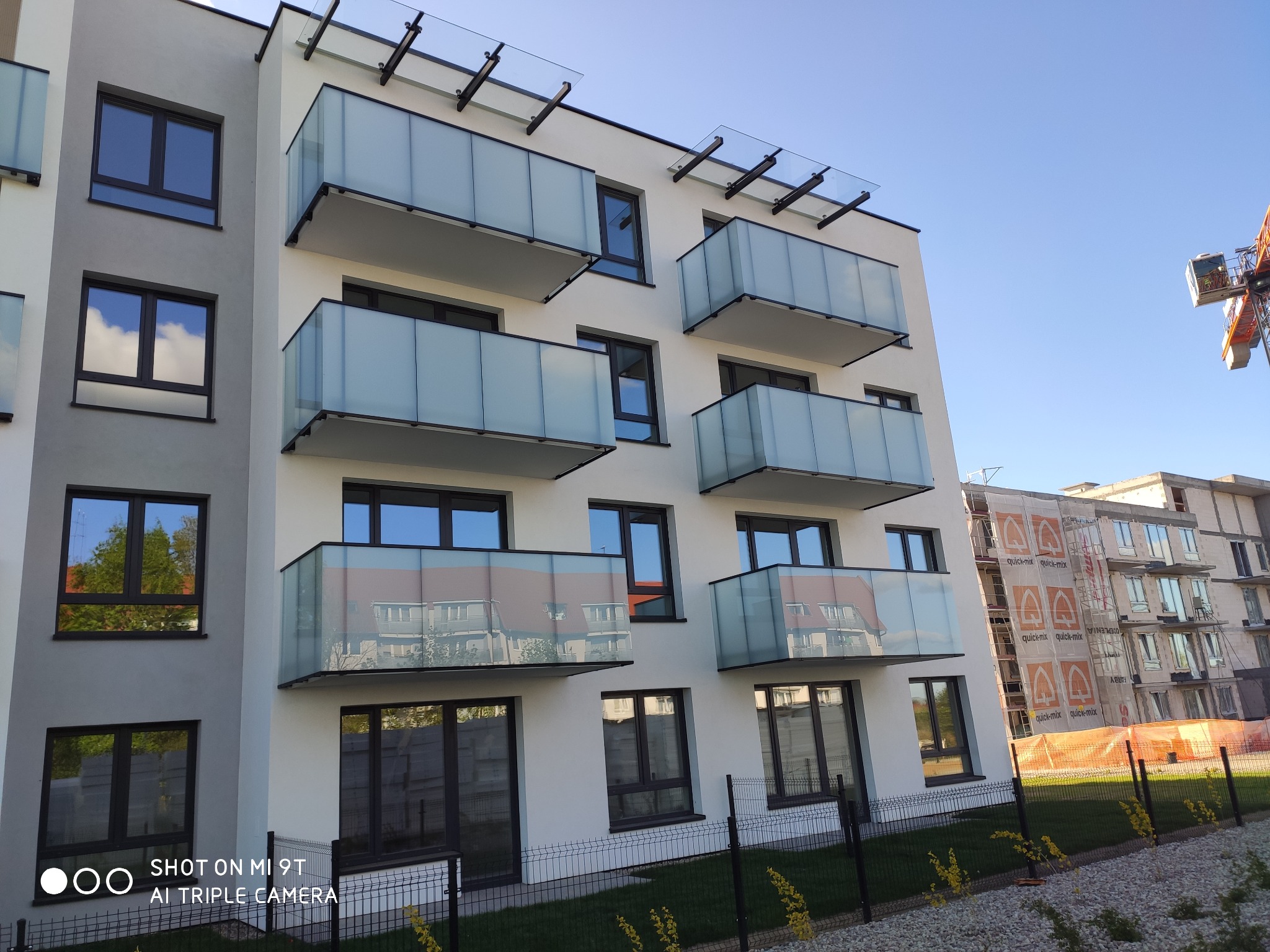 Nowoczesny budynek mieszkalny z balkonami z szklanymi balustradami i czarnymi ramami, widoczne odbicia w szybach, w tle budowa i żuraw.