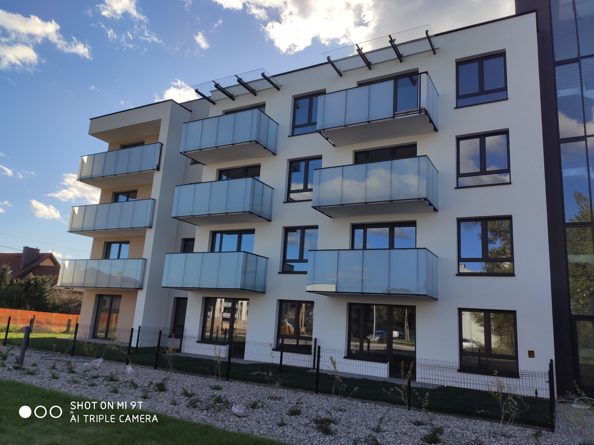 Nowoczesny budynek mieszkalny z balkonami z przezroczystymi szklanymi balustradami, widoczne mocowania balustrad do sufitu balkonów, błękitne niebo z chmurami odbija się w szybach okien.