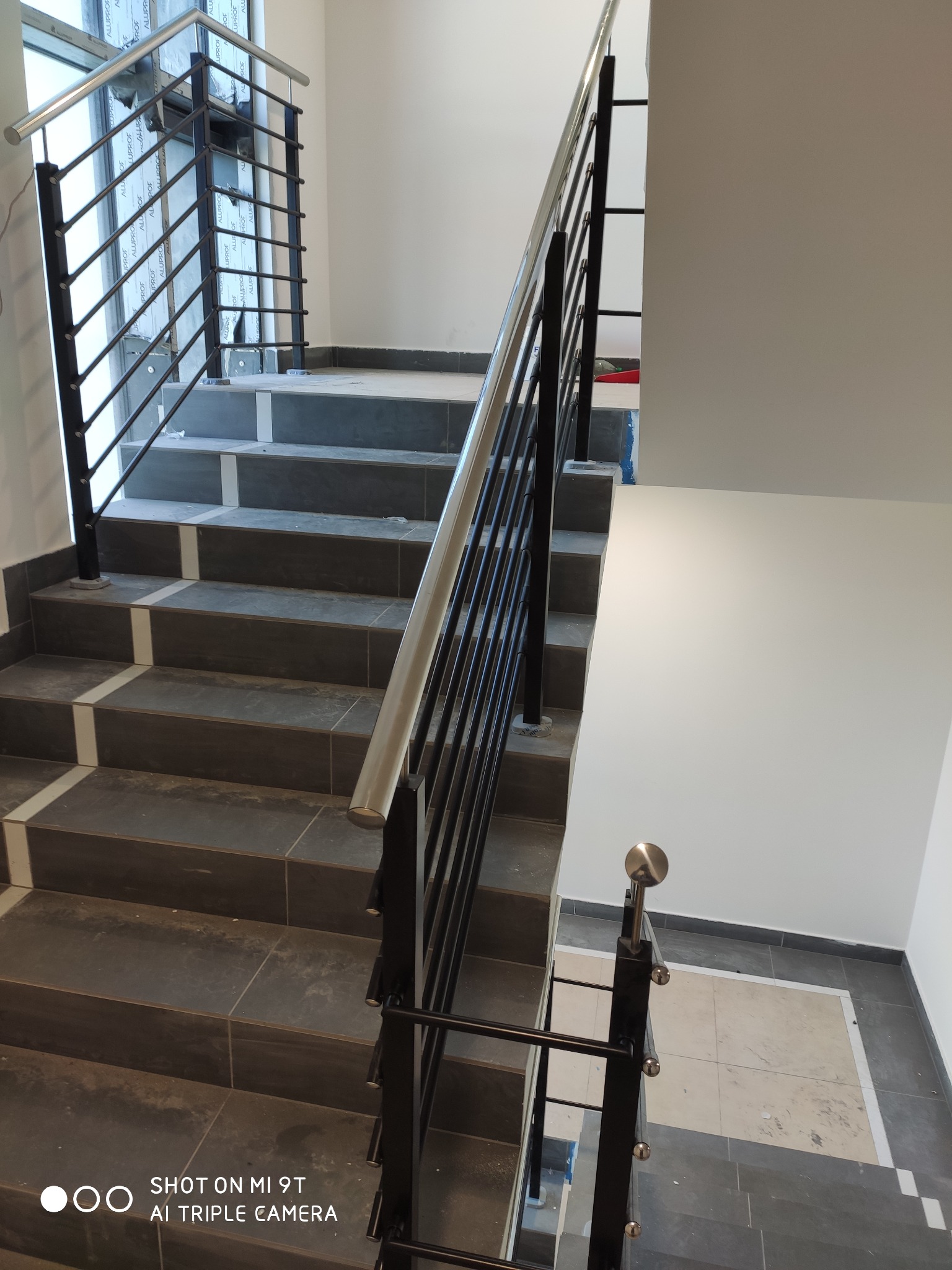 Schody wyłożone ciemnymi płytkami z jasnymi wstawkami, zabezpieczone czarną metalową balustradą z poziomymi szczebelkami i srebrną poręczą.