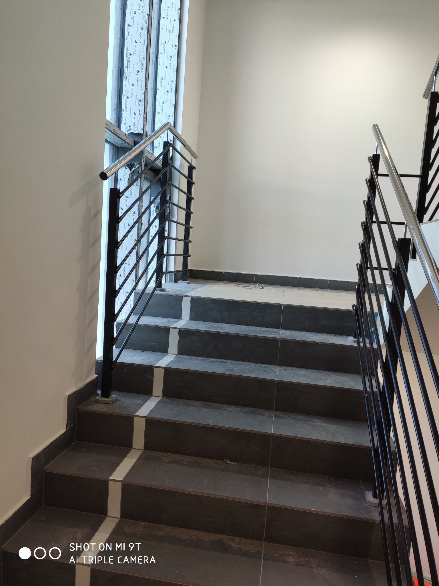 Schody wykończone szarymi płytkami z metalową poręczą z czarnymi pionowymi prętami i srebrną rurką na górze, widoczne okno z folią ochronną.