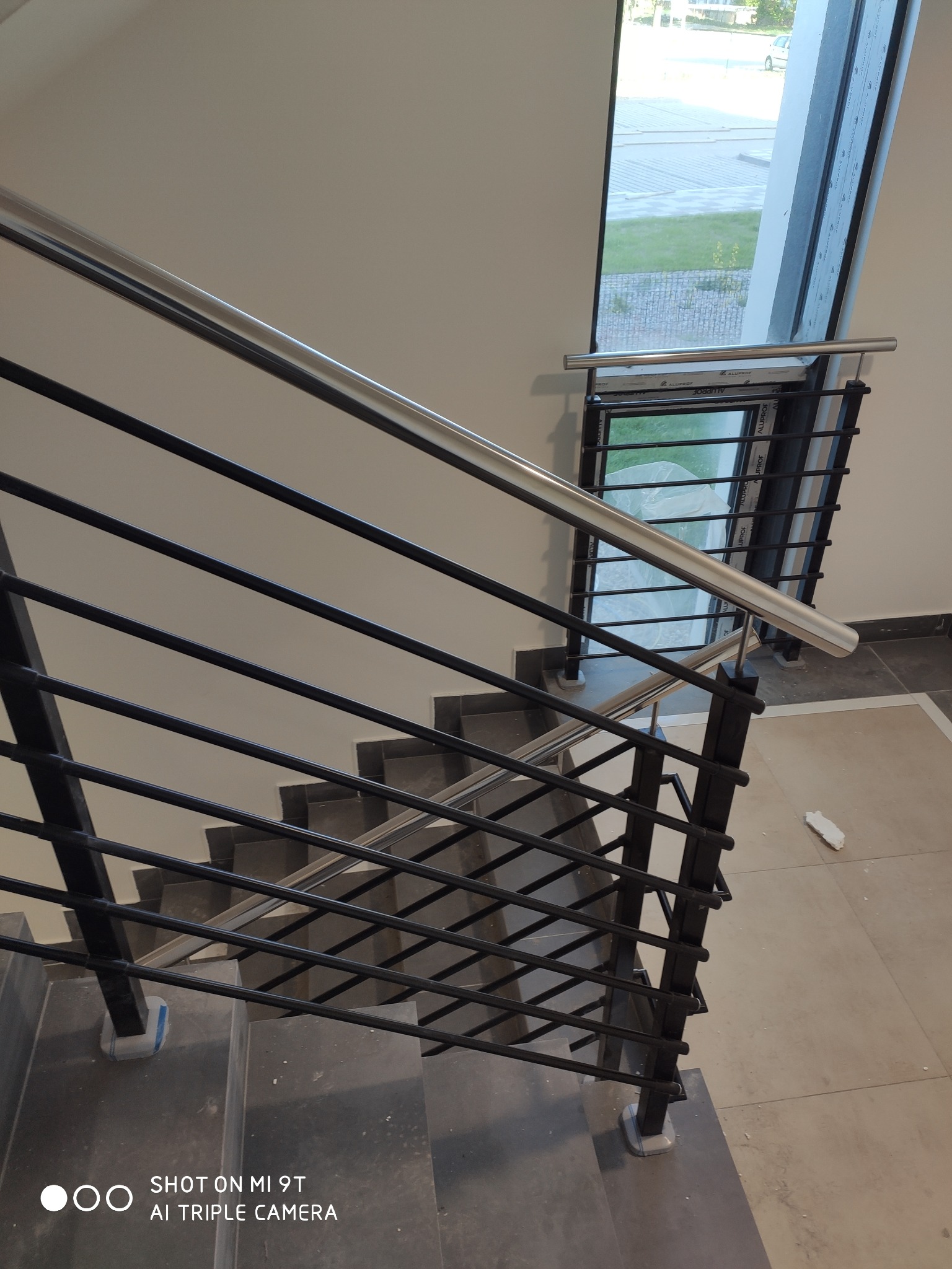 Wewnętrzna klatka schodowa z czarną balustradą z poziomymi prętami i nierdzewną poręczą, widok z góry na schody z szarymi płytkami, okno w tle.