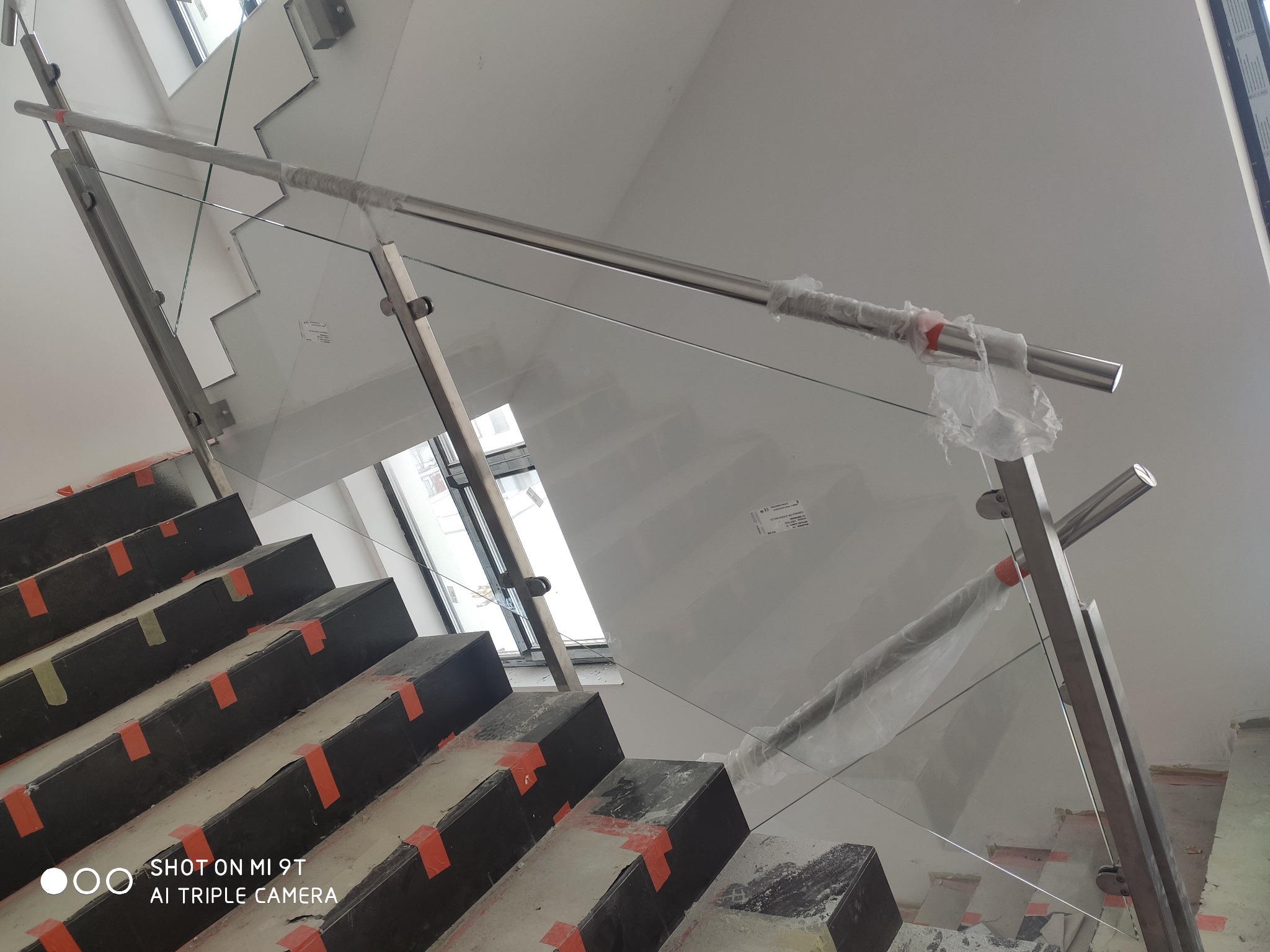 Montaż szklanej balustrady na schodach wewnętrznych, zabezpieczone folią ochronną, widoczne elementy montażowe.
