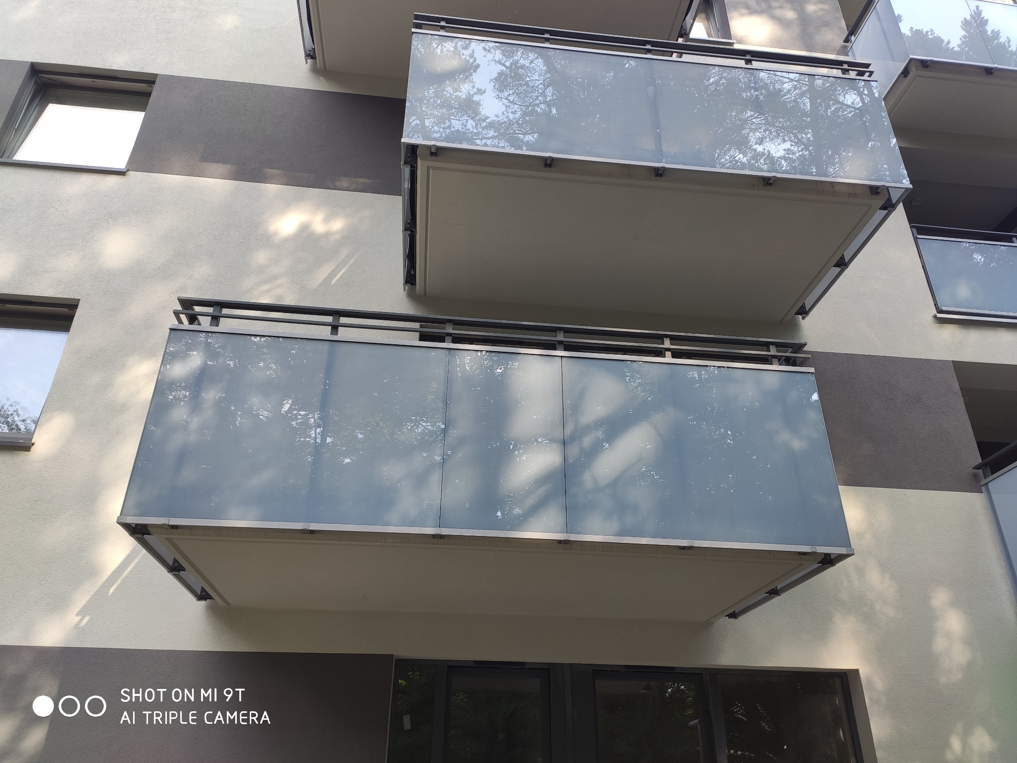 Zewnętrzny widok budynku z balkonami z szklanymi balustradami, odbijającymi korony drzew i niebo.