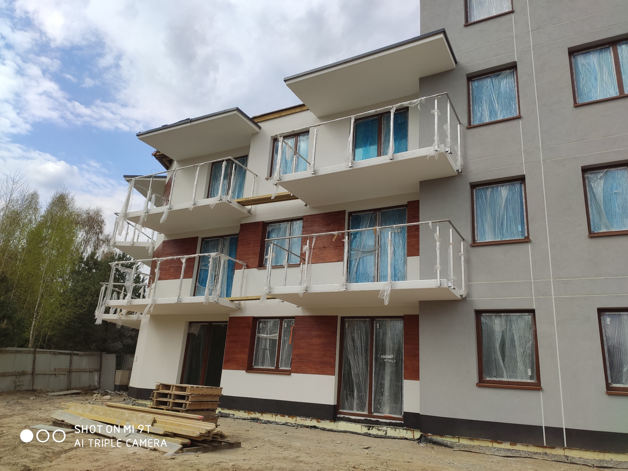 Nowoczesny budynek wielorodzinny w trakcie budowy z zamontowanymi białymi balkonami z metalowymi balustradami, okna zabezpieczone folią ochronną, widoczne materiały budowlane na placu budowy.