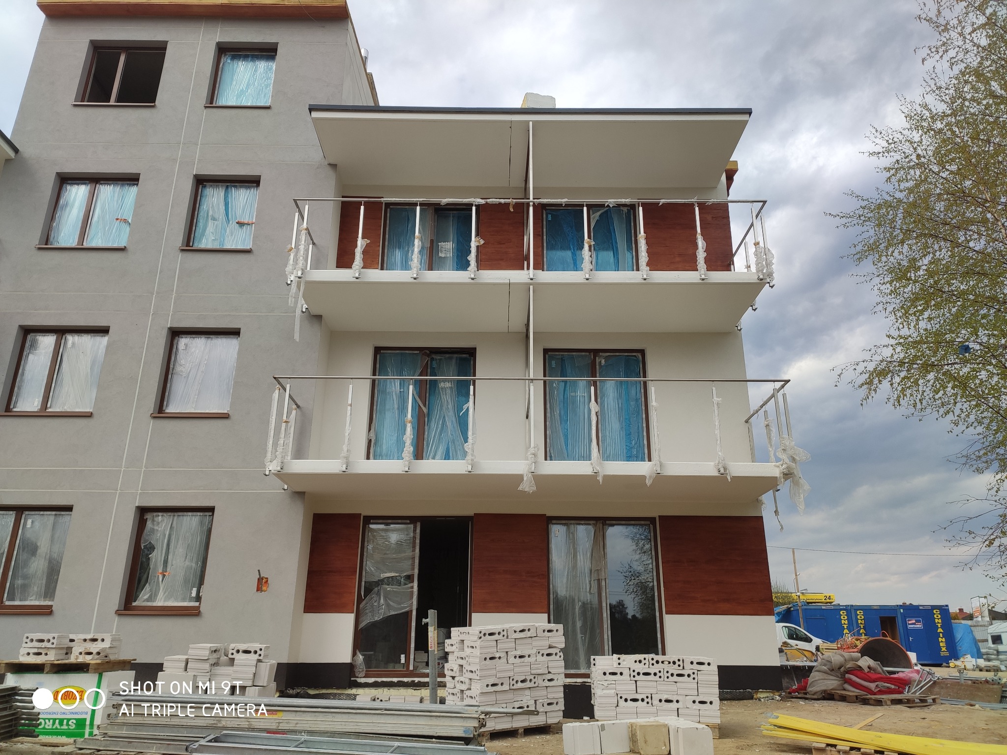 Budynek w Lidzbarku Warmińskim w trakcie budowy z zamontowanymi, zabezpieczonymi folią oknami i balustradami nierdzewnymi na balkonach, widoczne materiały budowlane.