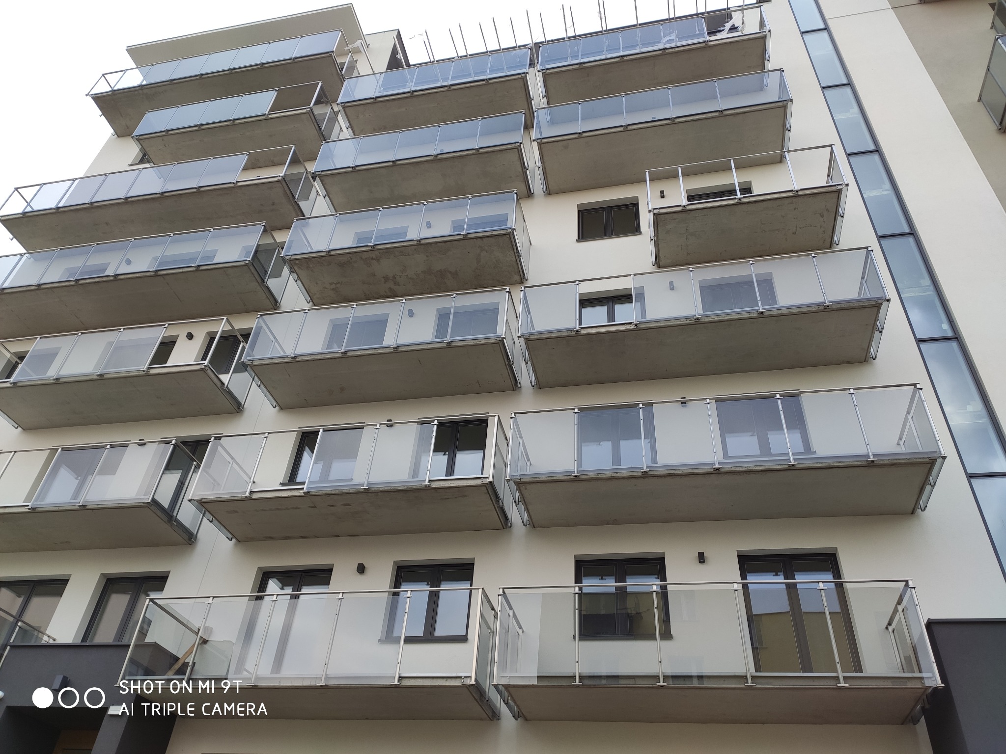 Fasada budynku wielorodzinnego z balkonami zabezpieczonymi szklanymi balustradami na metalowych słupkach, widok z dołu.