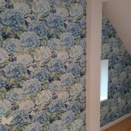 Ściana w pokoju pokryta tapetą w niebieskie hortensje, widoczny fragment okna i podłoga z paneli.