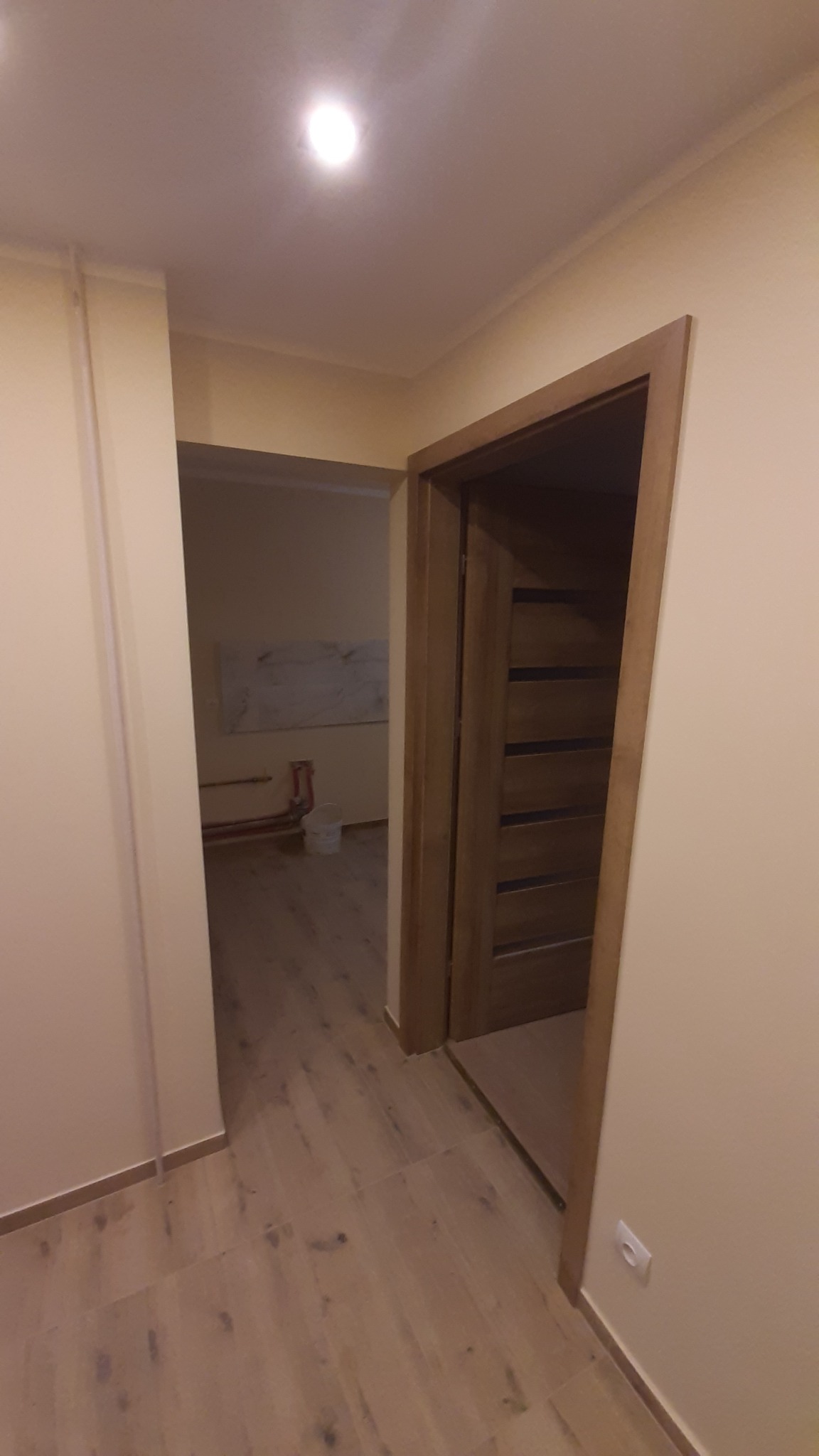 Wykończone wnętrze mieszkania po remoncie: drewnopodobne panele podłogowe, ościeżnica i drzwi z drewnianymi akcentami. Widoczne fragmenty instalacji w tle.