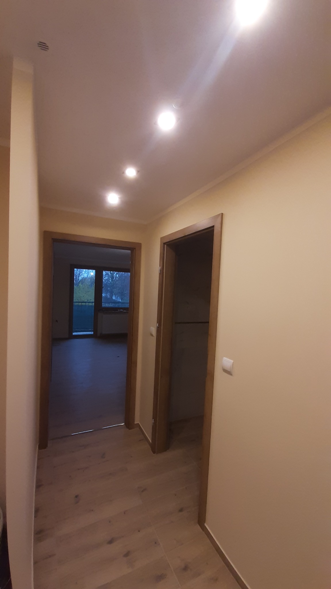 Jasny korytarz po remoncie z drewnianą podłogą i ościeżnicami. Widoczne drzwi do pokoi i okno z balkonem w tle. Oświetlenie sufitowe punktowe.