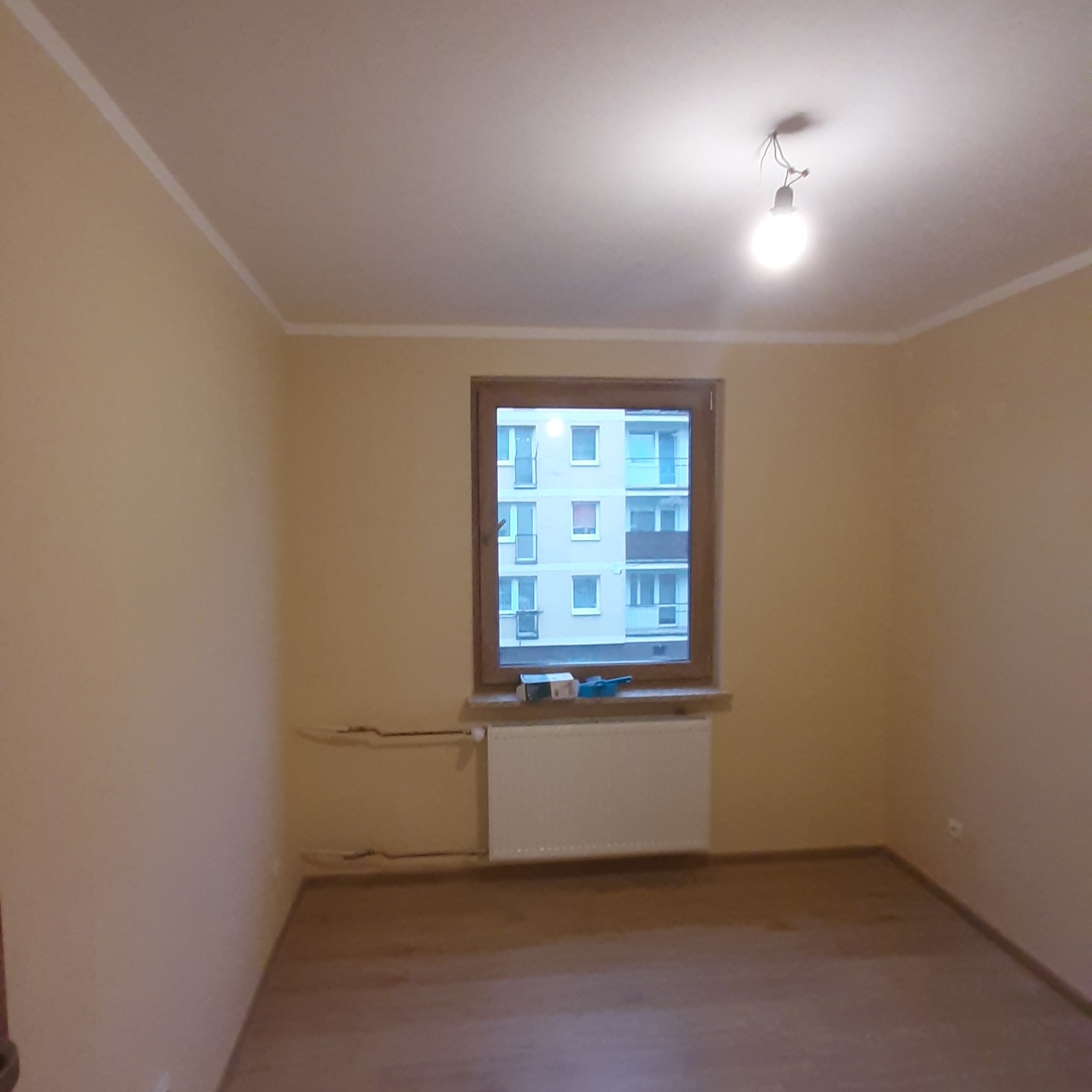 Pusty pokój z drewnianą podłogą i oknem z widokiem na blok. Ściany w odcieniach beżu, widoczny grzejnik pod oknem i wystające kable. Przygotowanie do prac wykończeniowych.