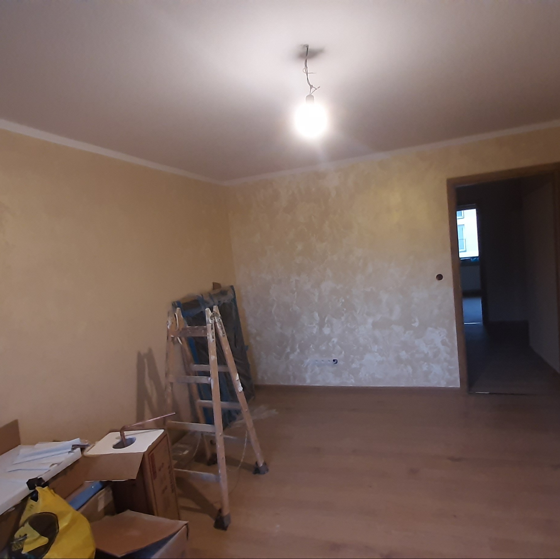 Pokój po malowaniu ścian z efektem dekoracyjnym, widoczna drabina malarska i kartony. Podłoga wyłożona panelami. Widok na otwarte drzwi do kolejnego pomieszczenia.