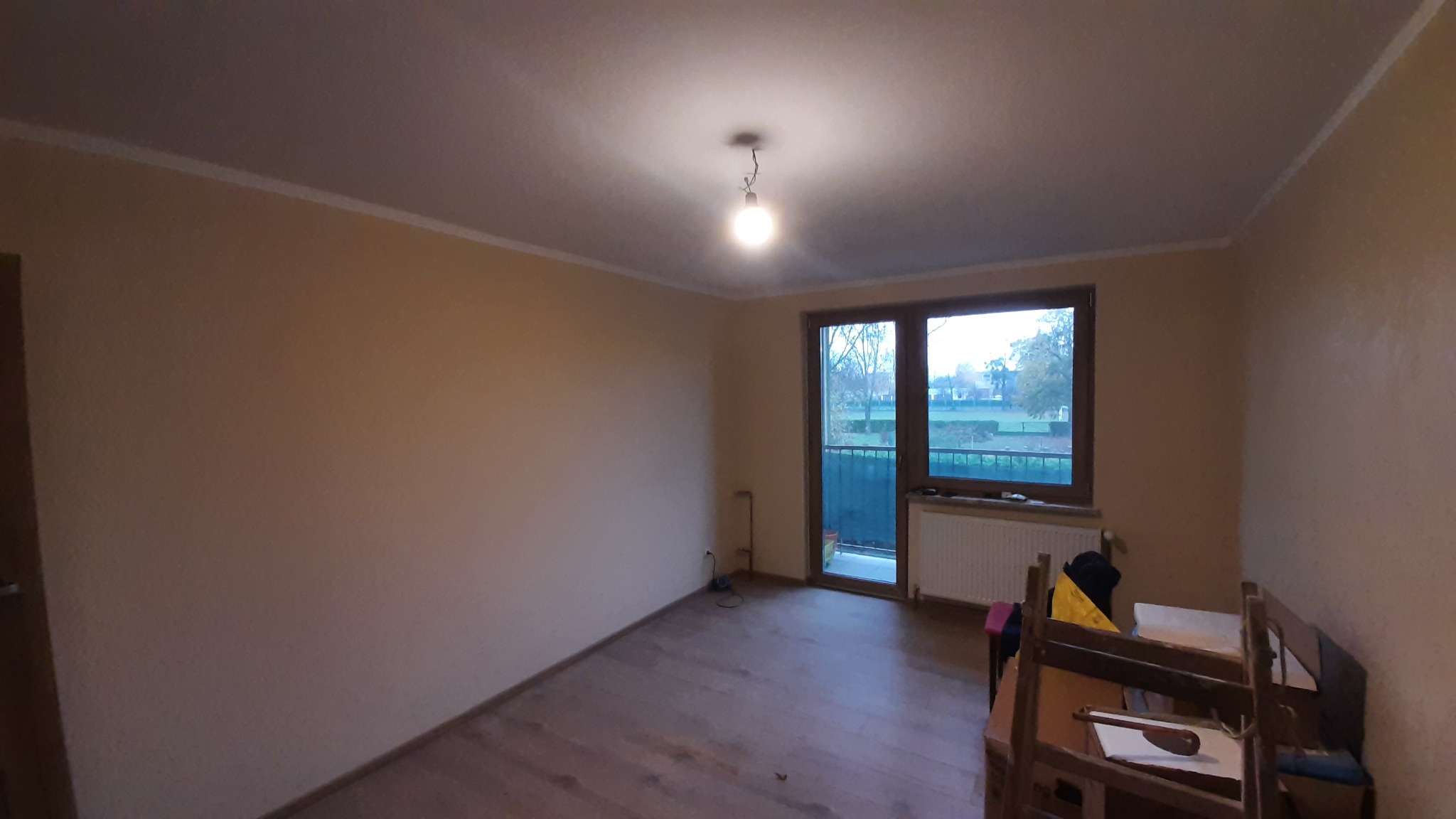 Pusty pokój po remoncie z drewnianą podłogą, beżowymi ścianami i balkonem. Widoczny grzejnik i lampa sufitowa. Przygotowany do umeblowania.