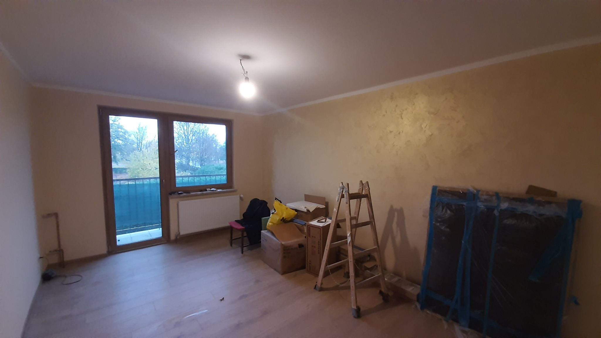 Pokój w trakcie prac wykończeniowych: drewniana podłoga, ściana z dekoracyjną fakturą, drabina malarska, okno balkonowe. Widoczne materiały budowlane.