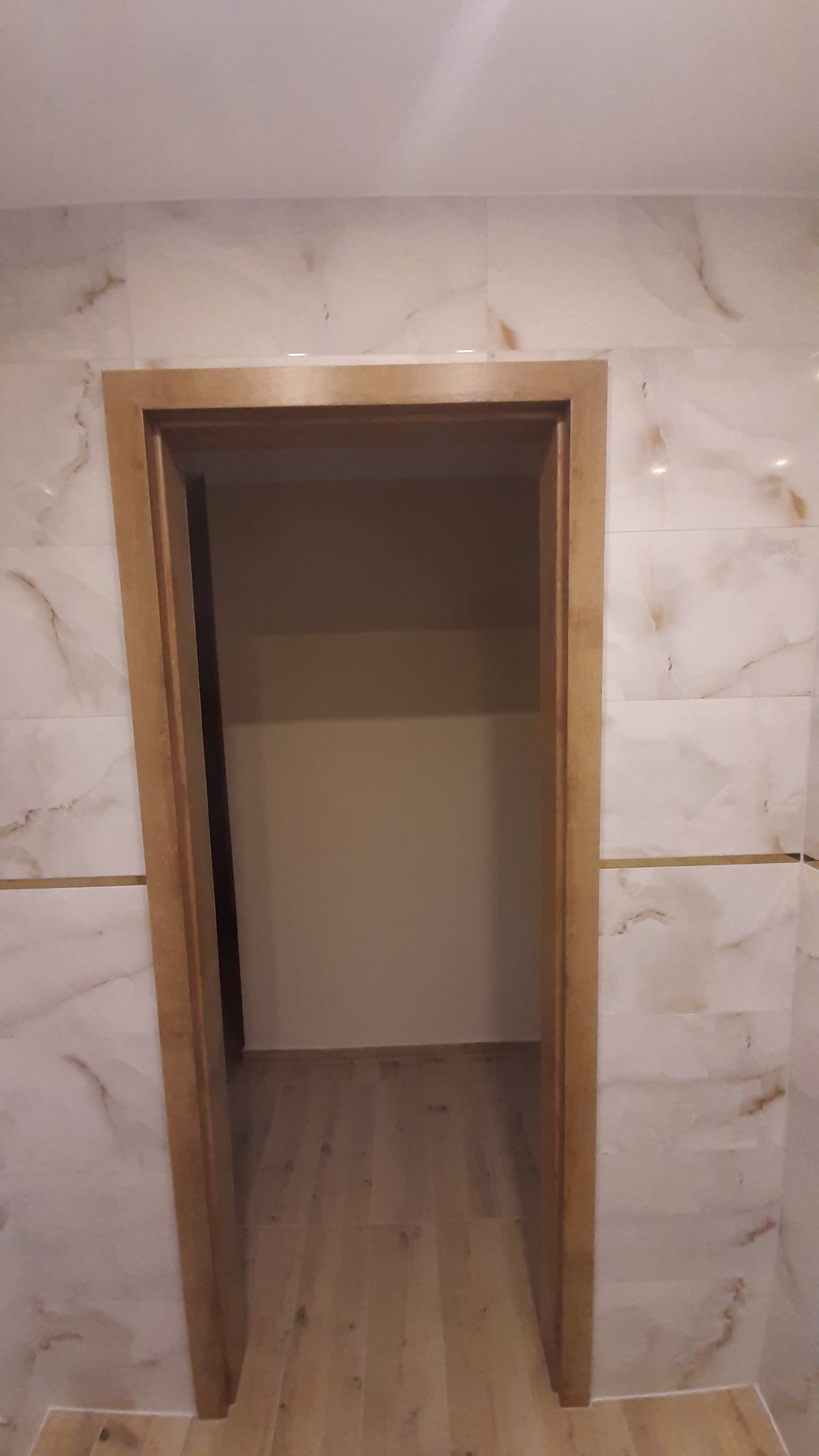 Wykończone wnętrze: marmurowe płytki ścienne z złotymi akcentami, drewniana ościeżnica i podłoga. Widok na sąsiednie pomieszczenie z beżową ścianą.