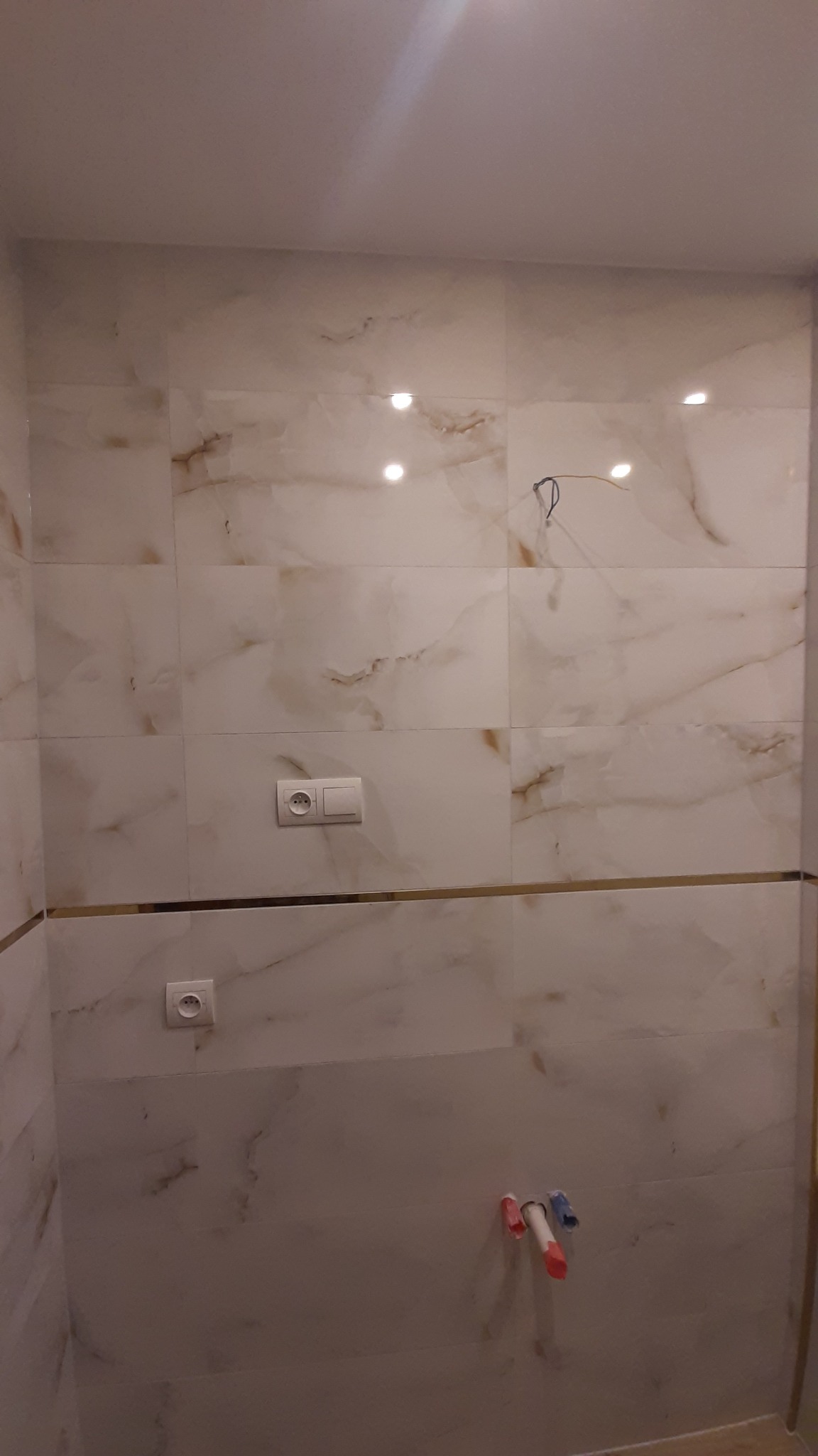 Wykończona ściana w łazience z marmurowymi płytkami, gniazdkami i instalacją wodną. Widoczna złota listwa dekoracyjna.