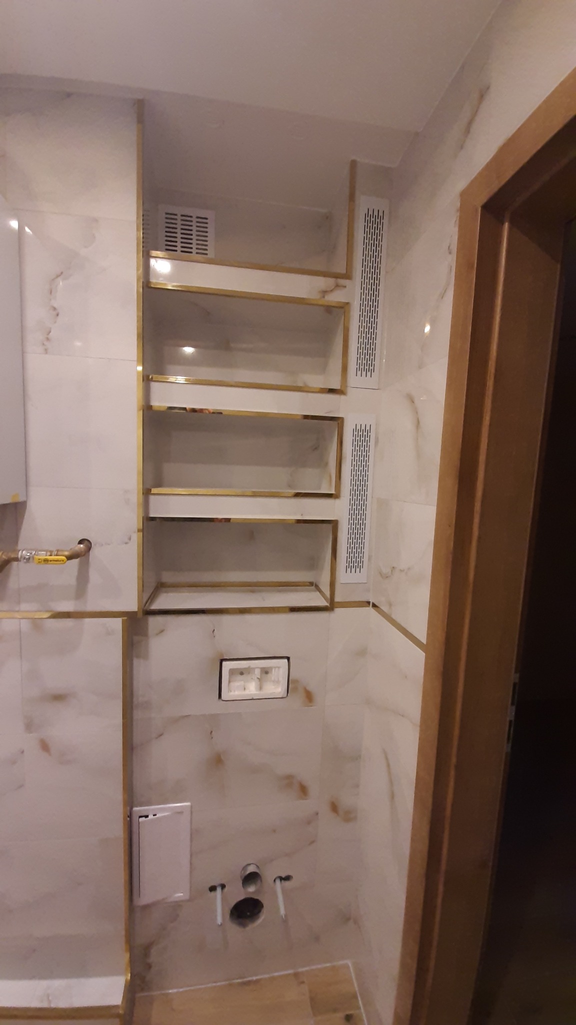 Wykończona łazienka z marmurowymi płytkami, złotymi akcentami i wnękami na półki. Widoczne przyłącza wodne i wentylacja. Drzwi z drewnianą ościeżnicą.