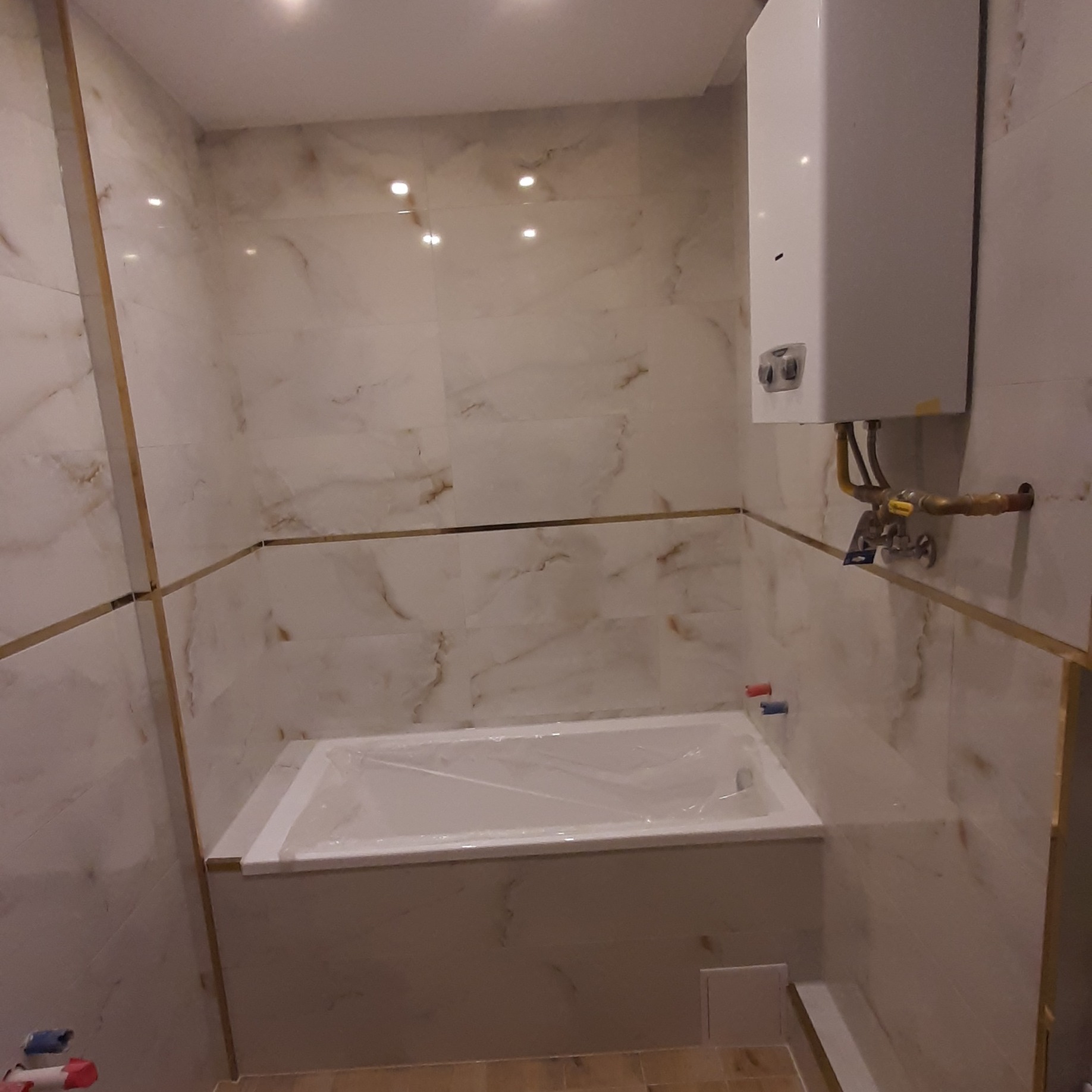 Wykończona łazienka w Kluczborku z wanną w folii ochronnej i marmurowymi płytkami z złotymi listwami. Biały piec gazowy na ścianie.