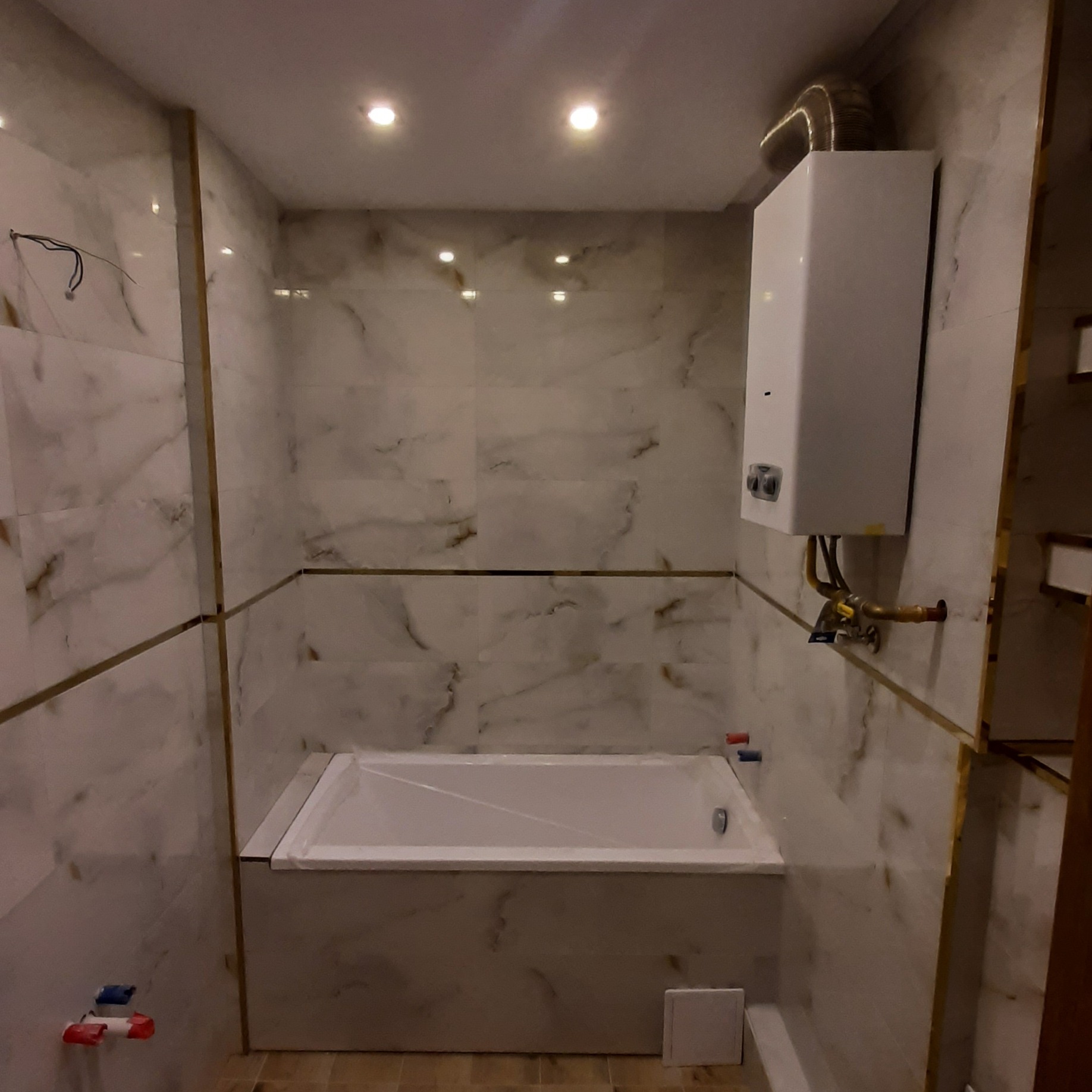 Wykończona łazienka z wanną obudowaną płytkami imitującymi marmur, z zamontowanym bojlerem i złotymi listwami dekoracyjnymi na ścianach.