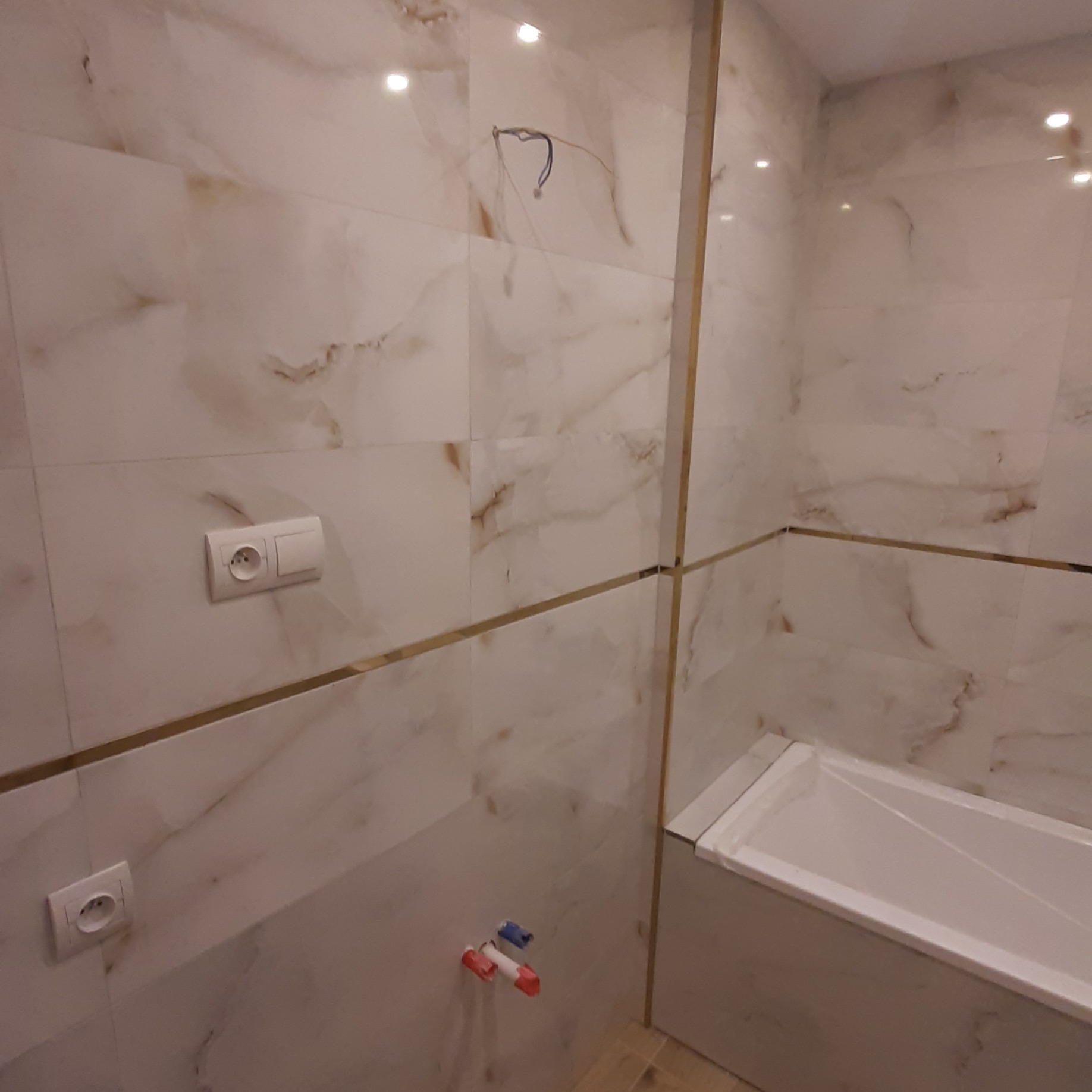 Wykończona łazienka z beżowymi płytkami imitującymi marmur, złotymi listwami, białą wanną i gniazdkami elektrycznymi. Widoczne przyłącza wodne.